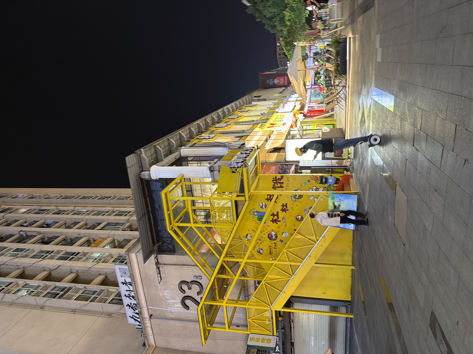 This image depicts an outdoor street scene at night, likely in the Chongqing High-tech Zone, China, characterized by a tall, modern building and a brightly lit commercial area.
Dominating the left side of the frame is a tall, multi-story building with numerous windows and a neutral-colored facade. Below this building, a prominent, vibrant yellow, multi-level outdoor staircase structure is a focal point. This structure features metal railings and a glass-enclosed top section. The yellow staircase is adorned with cartoonish illustrations and text, including "HAVE FUN" and Chinese characters.
In front of the yellow staircase, two individuals are present. One person, wearing a light-colored shirt and dark pants, stands near the bottom of the stairs, looking at a phone. Another person, dressed in a dark jacket and jeans, is actively riding a self-balancing scooter (hoverboard) on the paved walkway in the mid-ground. This individual is also looking down, possibly at a device.
The walkway is paved with grey rectangular tiles. To the right of the yellow staircase, a row of shops or restaurants is visible, with illuminated signs and awnings. Several outdoor tables and chairs are set up under umbrellas, suggesting a dining or leisure area. The background shows more buildings and trees, illuminated by artificial lights, contributing to the lively night atmosphere of an urban commercial district. The overall lighting is artificial, casting a warm glow on the scene and creating reflections on the wet pavement.