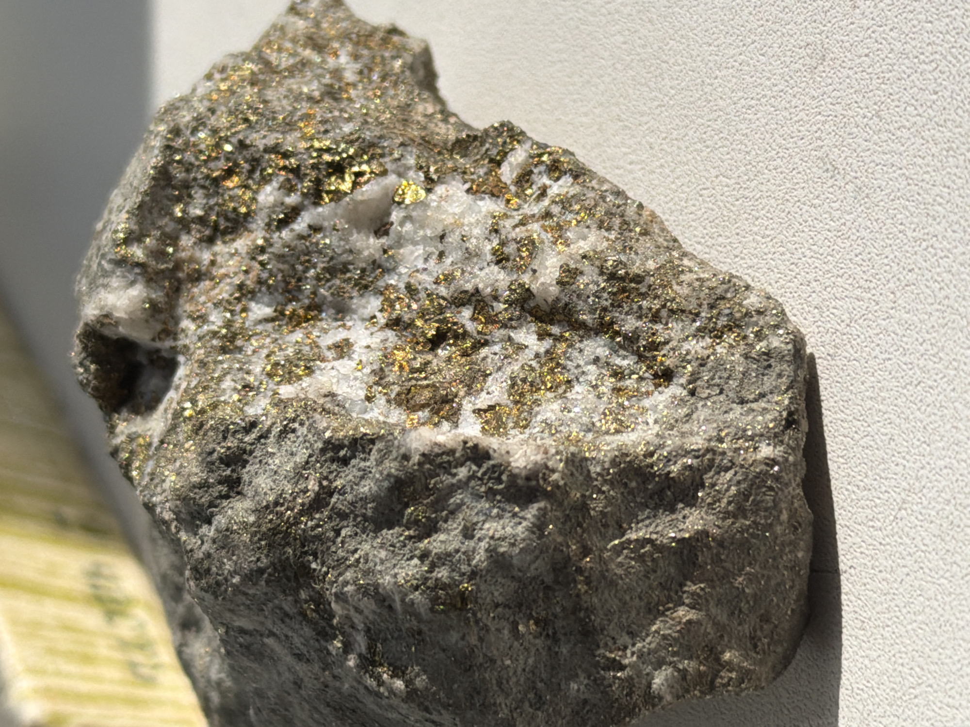 A close-up shot of a rock specimen, predominantly gray and textured, with visible specks of gold-colored metallic minerals. The rock also contains a section of white, crystalline material. The background is a solid, light-colored surface, possibly a wall. The image does not contain any information that would allow one to determine the location as Izhevsk, Russia, or any information about people, actions, or interactions.