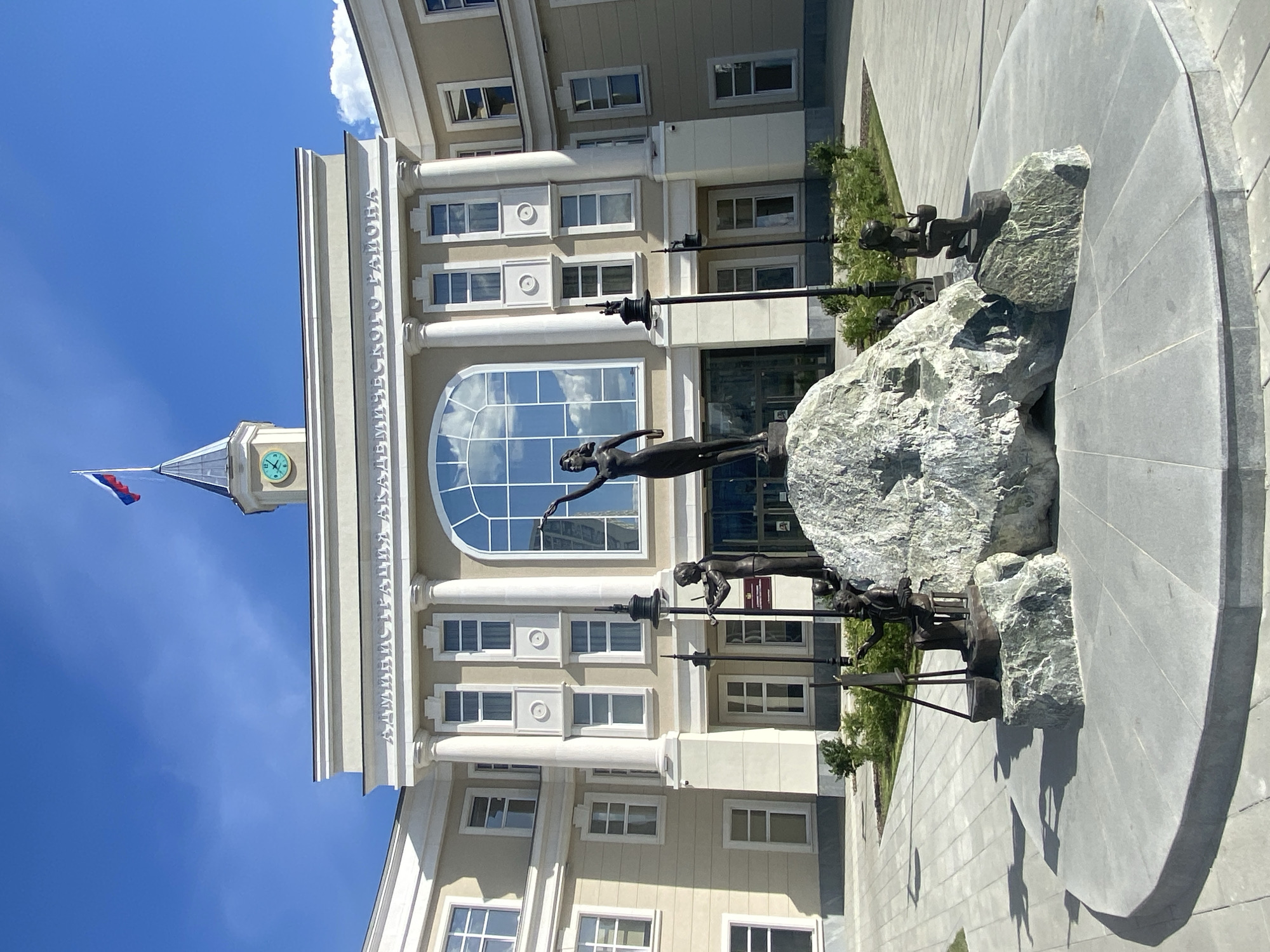 The image depicts a building with a monument in front of it, located in Yekaterinburg, Russia. The building is beige with white columns and a large arched window reflecting the sky. Above the window is the inscription "Администрация Академического района". A tower with a clock and a Russian flag extends from the left side of the building. In front of the building is a monument consisting of a group of bronze sculptures standing atop large rocks. The monument sits on a circular gray stone platform. The sky is clear and blue.