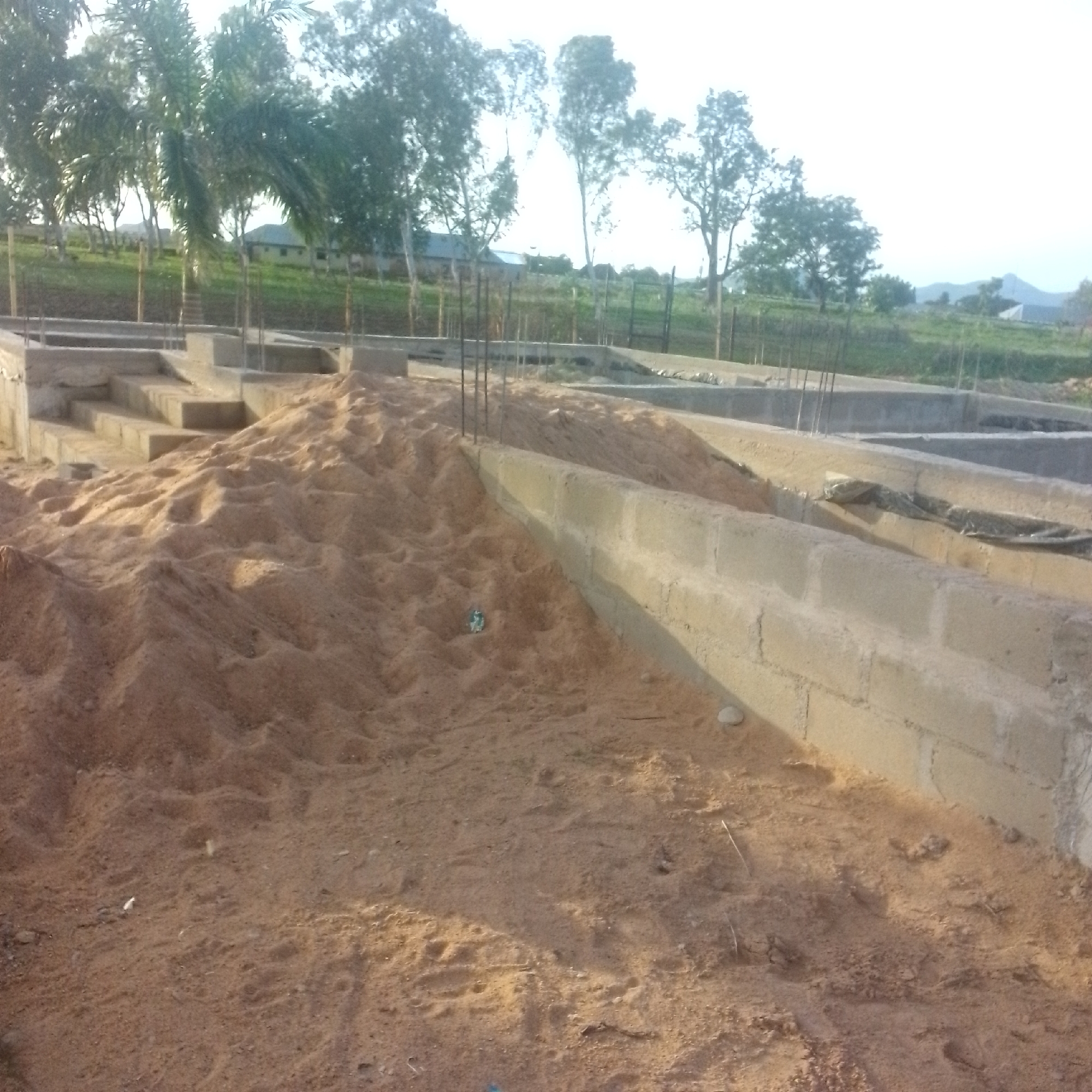 The image depicts the foundation of a building under construction in Jalingo, Nigeria. The foreground is dominated by a large pile of sand, with some visible rebar sticking out. To the left, concrete steps lead up to a higher level of the foundation. A concrete block wall forms the side of the foundation, with more rebar protruding. In the background, several trees, including palm trees, are visible, suggesting a tropical or sub-tropical environment. Beyond the trees, there are some buildings, possibly houses or other structures. The sky is bright and overcast, indicating it is daytime, likely morning or late afternoon due to the lighting. The overall scene suggests an active construction site. There is no visible text in the image.