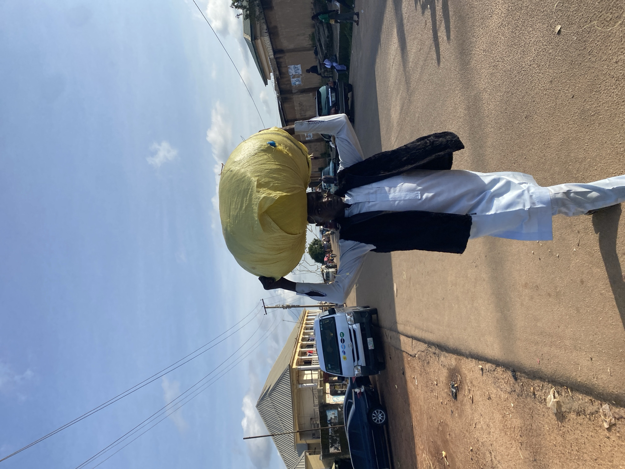 A man is carrying a large, yellow, round object on his head as he walks down a street in Gura Top, Nigeria. He is wearing a white traditional outfit and a dark vest. The street is paved and has a dirt shoulder. There are buildings and parked cars on either side of the street, as well as utility poles and power lines. The sky is blue with some scattered clouds. The man is facing the camera and appears to be walking towards the viewer. The overall scene suggests a typical day in a busy street market environment.