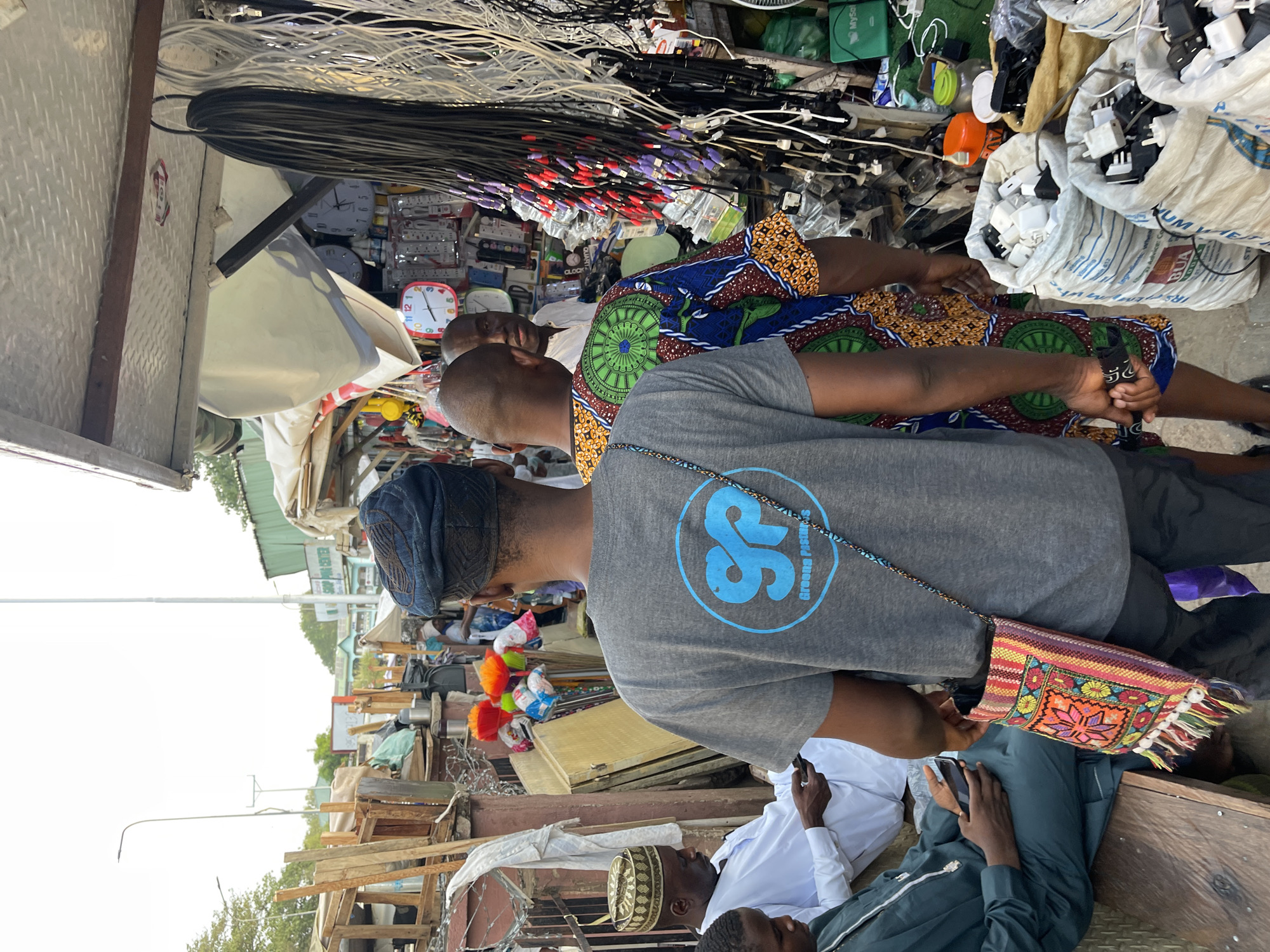 This image captures a vibrant market scene in Abuja, Nigeria, during daylight hours. The setting appears to be a roadside stall or a section of a larger market, characterized by various goods displayed and multiple individuals.
In the foreground, several people are present. A man wearing a grey t-shirt with a blue "SS Greena Pastures" logo is central, viewed from behind. He has a colorful, patterned woven bag with tassels slung over his shoulder. Next to him, another man wears a bright, patterned shirt, likely of traditional West African fabric. Further back, individuals are seen in white and dark traditional attire, some wearing kufis or similar head coverings. One person is seen looking at a mobile phone.
The market stall is filled with a dense array of merchandise, primarily electronics and household items. Prominently featured are bundles of electrical cables, including black and white varieties with various connectors such such as RCA and VGA. Bags filled with white and black electrical adapters or chargers are also visible. Among the other goods are multiple wall clocks, some round and some square, displaying numbers like "1, 3, 6, 9, 10, 11, 12" and the words "CLOCK" and "QUARTZ." Other small electronics and items are piled around. A green bag with "MySol" written on it is discernible in the background, alongside white bags that have "BUA" and "UM WE" printed on them. The overall impression is one of a busy, functional marketplace offering various goods.