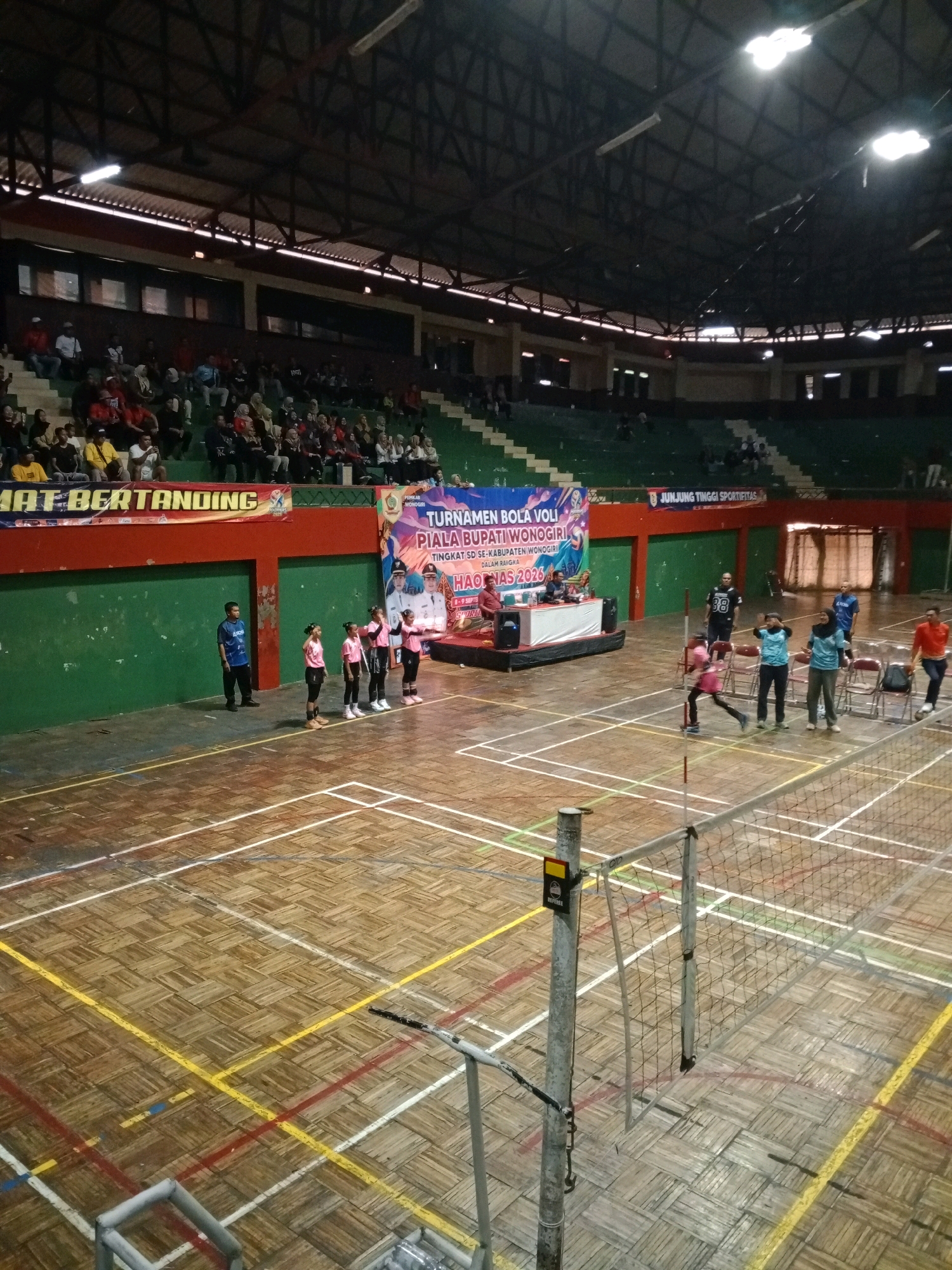 This image captures an indoor volleyball tournament at the GOR Wonogiri sports hall in Wuryorejo, Indonesia. The scene depicts a volleyball match in progress, with players on the court and spectators in the stands. The sports hall features a wooden floor marked with volleyball court lines, a net dividing the playing area, and bleacher seating surrounding the court. A banner in the background announces "TURNAMEN BOLA VOLI PIALA BUPATI WONOGIRI TINGKAT SD SE-KABUPATEN WONOGIRI DALAM RANGKA HAORNAS 2023," indicating the event is the Wonogiri Regent's Cup Volleyball Tournament for elementary schools, held in conjunction with National Sports Day (HAORNAS) 2023. Several individuals, including young girls in pink uniforms, appear to be participating in the tournament, while other people, presumably officials and spectators, are present around the court and in the stands. The lighting suggests it is daytime, though the indoor setting limits natural light.