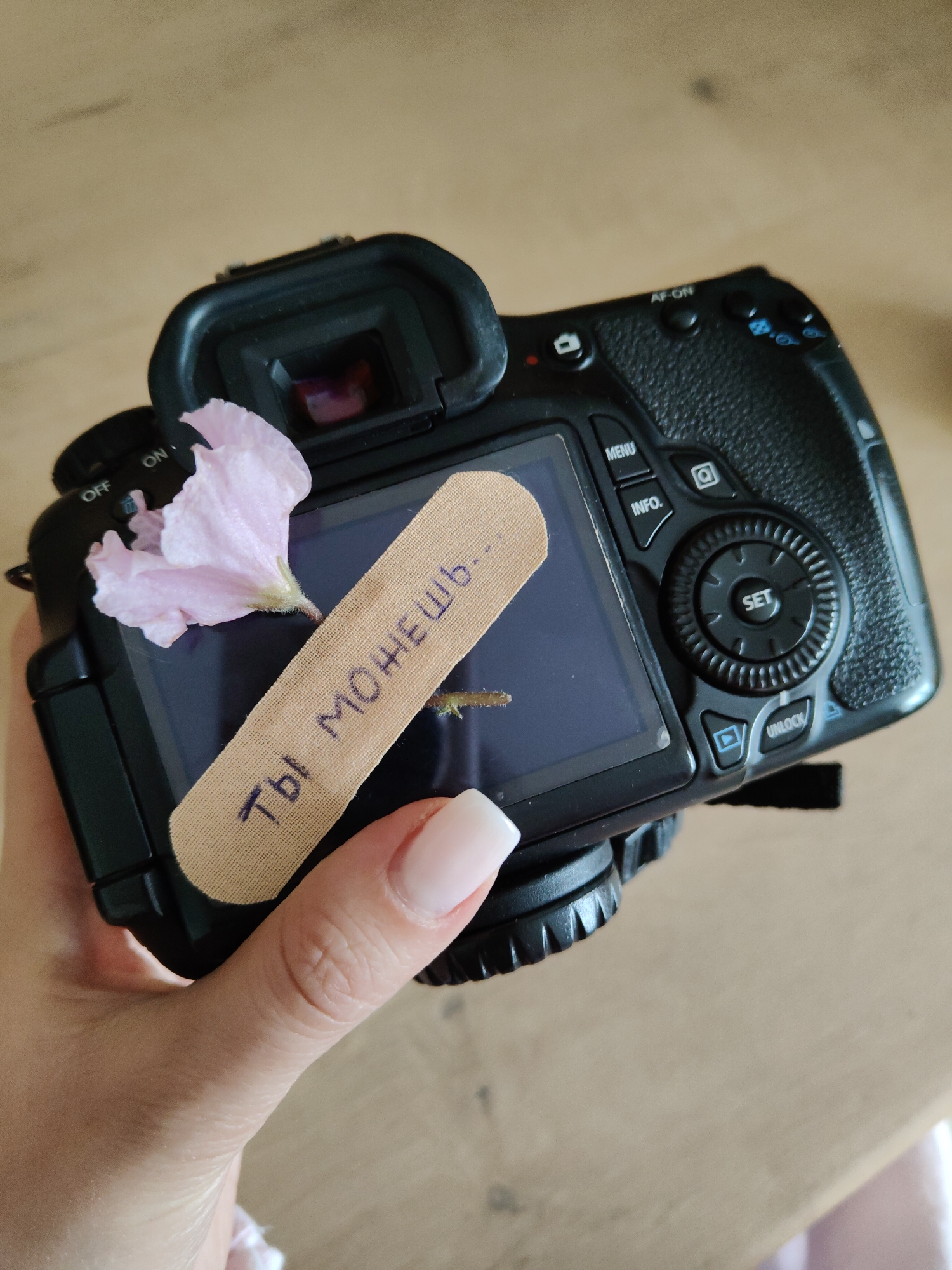 A person is holding a black digital camera in their hand. A pink flower is placed on the camera, partially covering the screen. A bandage with text written on it in what appears to be Cyrillic script covers the majority of the screen. The camera and the hand holding it appear to be on a wooden surface. The location is unknown.