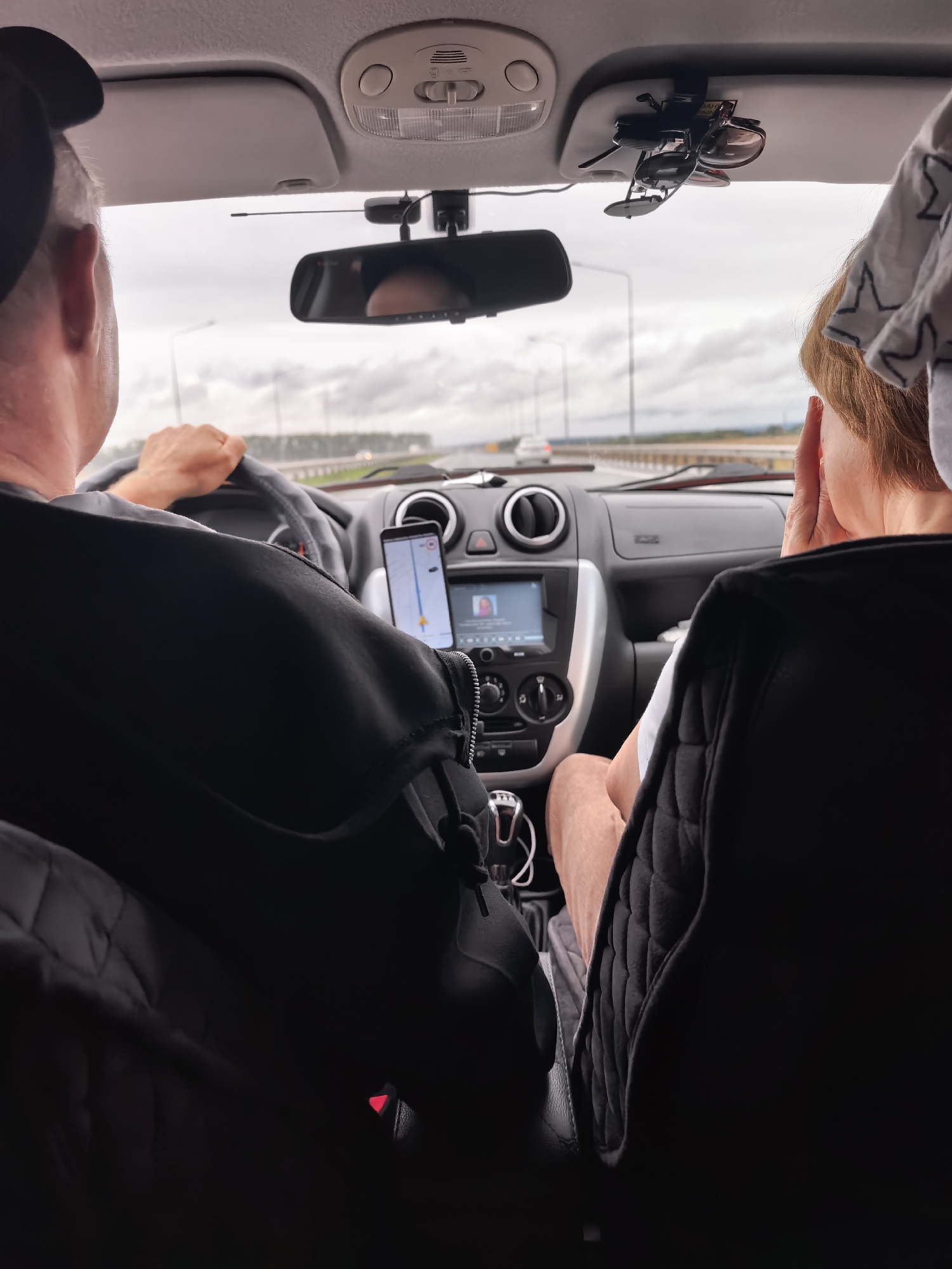 The image is a point-of-view shot from inside a vehicle traveling on a highway, likely near Dubki, Russia, given the context. A person wearing a black baseball cap and a black jacket is driving, with their hands on the steering wheel. To their right, another person, partially visible from behind, is sitting in the passenger seat with their hand on their face, appearing to be looking out the window. The car's interior shows a navigation system mounted on the dashboard with a map displayed, along with air vents and other controls. Sunglasses are clipped to the visor above. The road outside is overcast, with trees lining the median and other vehicles visible in the distance. The overall scene depicts a journey in progress.