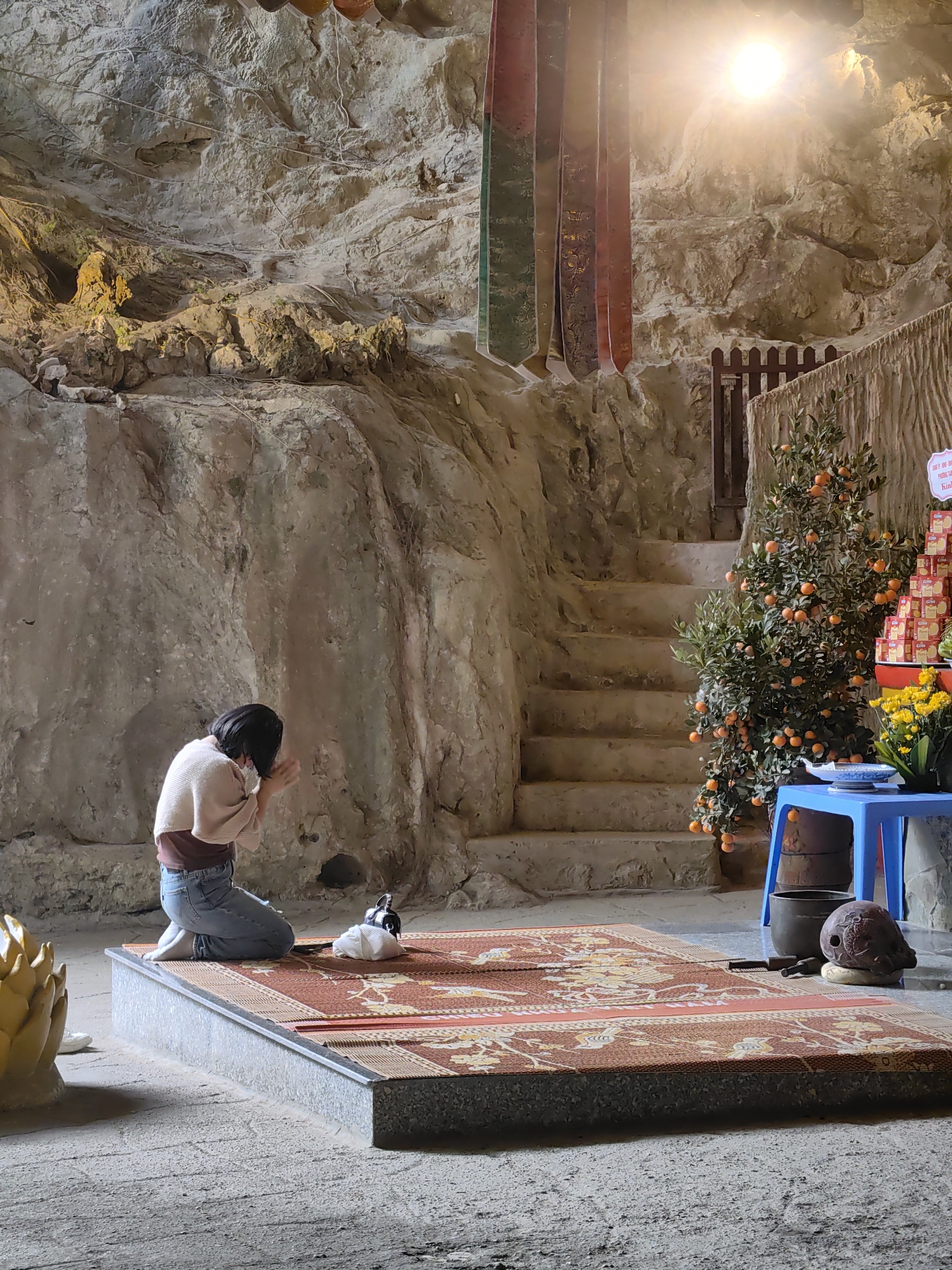 A full shot captures a person kneeling in prayer on a decorated platform within a cave-like structure. The individual is seen from the rear, wearing jeans and a tan top, hands clasped in front of their face. A white object, possibly a pet, sits nearby on the rug-covered platform. The cave's walls are rough and appear to be natural stone. Behind the platform, stairs ascend towards a wooden fence, with a small tree bearing orange fruits next to the stairs. To the right, a blue table is visible, with a stack of decorative boxes and yellow flowers. Above, sunlight streams in, illuminating the scene. Banners of varying colors hang from the ceiling. The picture was taken in Lạng Sơn, Vietnam.