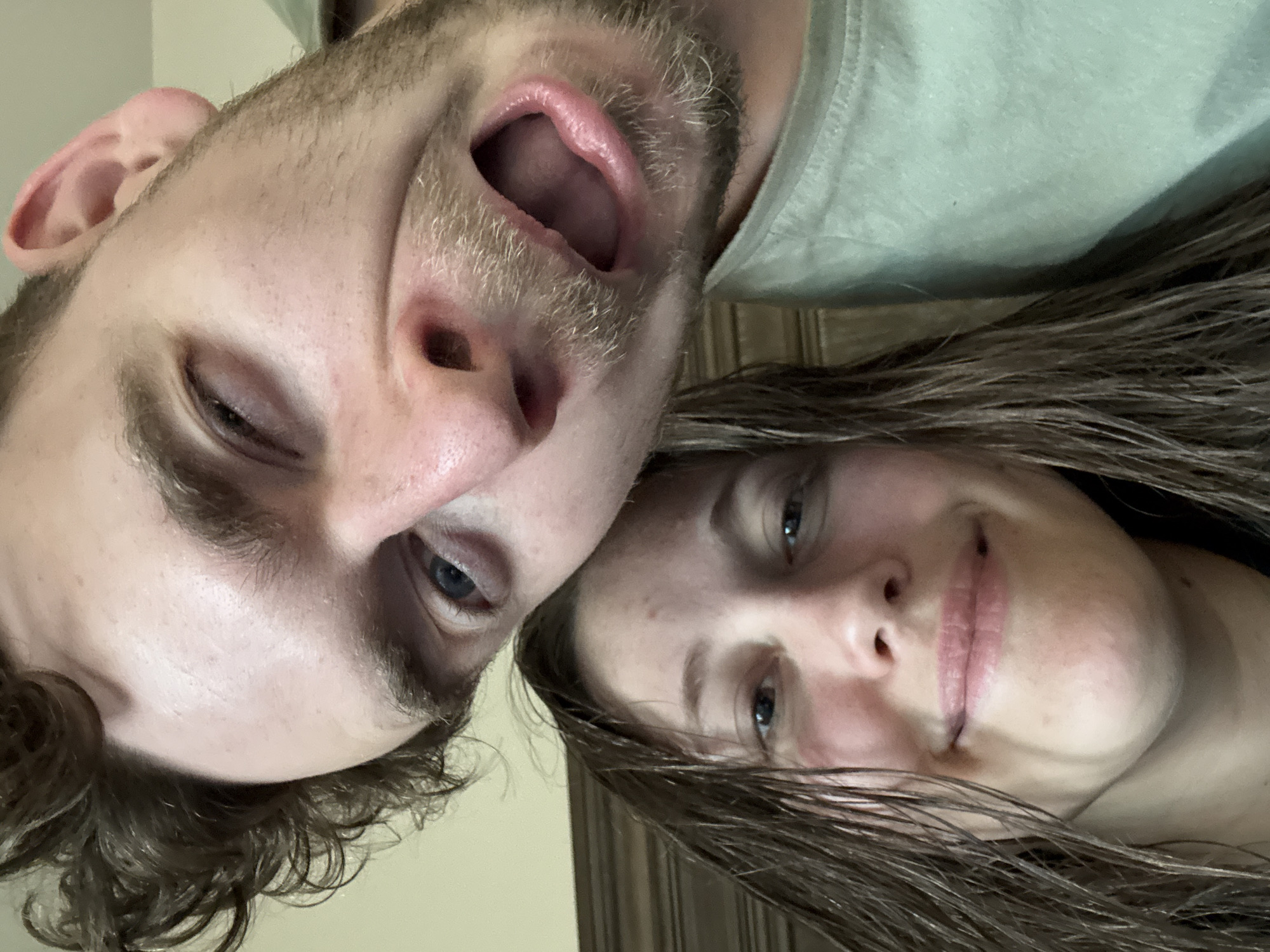 In a close-up selfie, a man and a woman are depicted. The man is positioned in the foreground, his face angled towards the camera with a playful, exaggerated expression, mouth open and one eye squinting. He has brown hair and facial hair. The woman is partially visible behind him, her head tilted and her face turned slightly towards the camera. She is smiling, and her brown hair appears wet. They are both looking at the camera. The background shows a wooden surface, likely furniture, and a light-colored wall. The image appears to have been taken indoors.