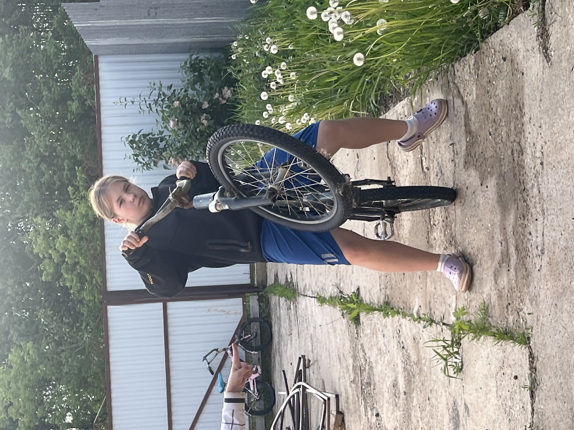 A young person stands in an outdoor location in what is assessed to be Chaikovsky, Russia. The individual is holding a bicycle, with the front wheel suspended in the air. The person is wearing a dark-colored jacket, blue shorts, and light purple slip-on shoes. The environment includes a metal fence, various bicycles, and green vegetation, including dandelion seed heads. The ground is concrete with weeds growing through the cracks. There is another person's hand visible in the lower left corner.