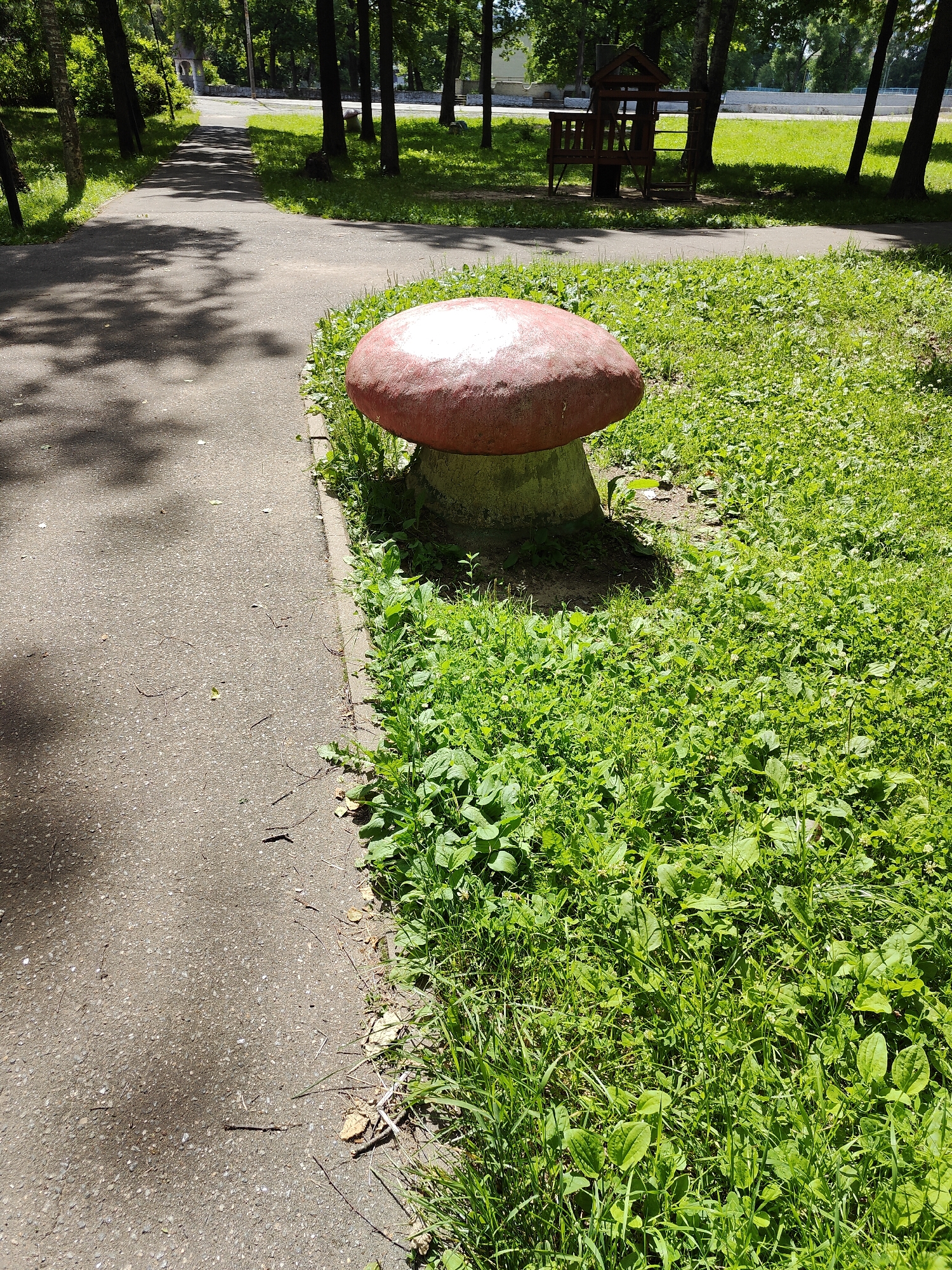 In an outdoor park setting, a large, decorative mushroom sculpture sits on a concrete base next to a paved walkway. The mushroom has a red cap with white speckles and a gray stem. The walkway is shaded by trees casting dappled shadows. To the right of the walkway, lush green grass and various plants cover the ground. In the background, a wooden playground structure is visible amongst trees, and beyond that, a white fence and a distant structure suggest the edge of the park. The scene is illuminated by bright daylight, indicating it is daytime. There are no people or animals visible in the image.