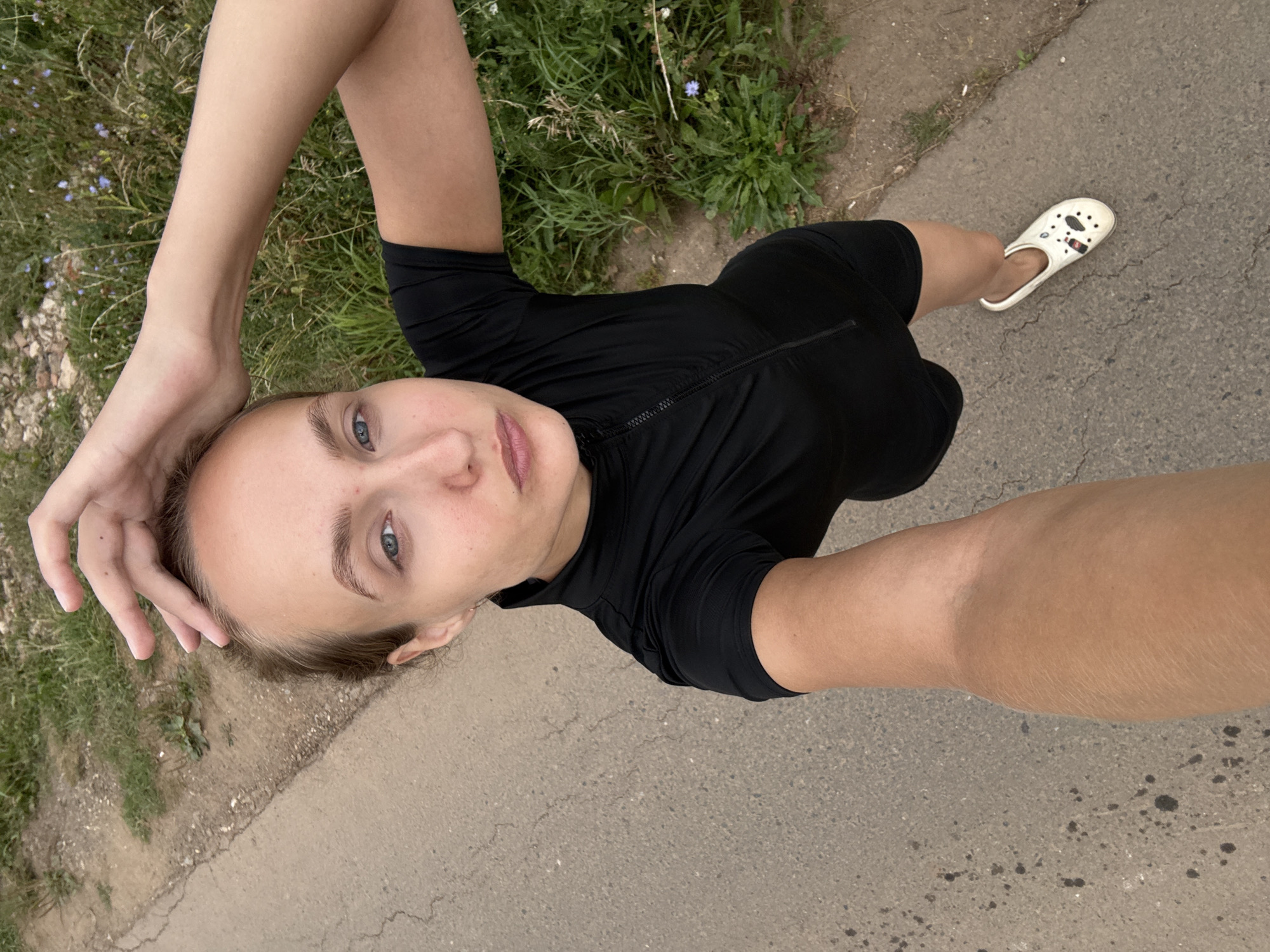 A woman is captured in a high-angle selfie from the perspective of her left arm and leg. She is standing on a paved surface adjacent to a grassy area with some wildflowers. The woman is wearing a black athletic top with a zipper and black shorts, and white Crocs with cartoonish designs. Her right hand is raised to touch the top of her head. She is looking directly at the camera with a pout. The lighting suggests it is daytime. The setting appears to be a rural road or path in or near Podshivalovo, Russia.