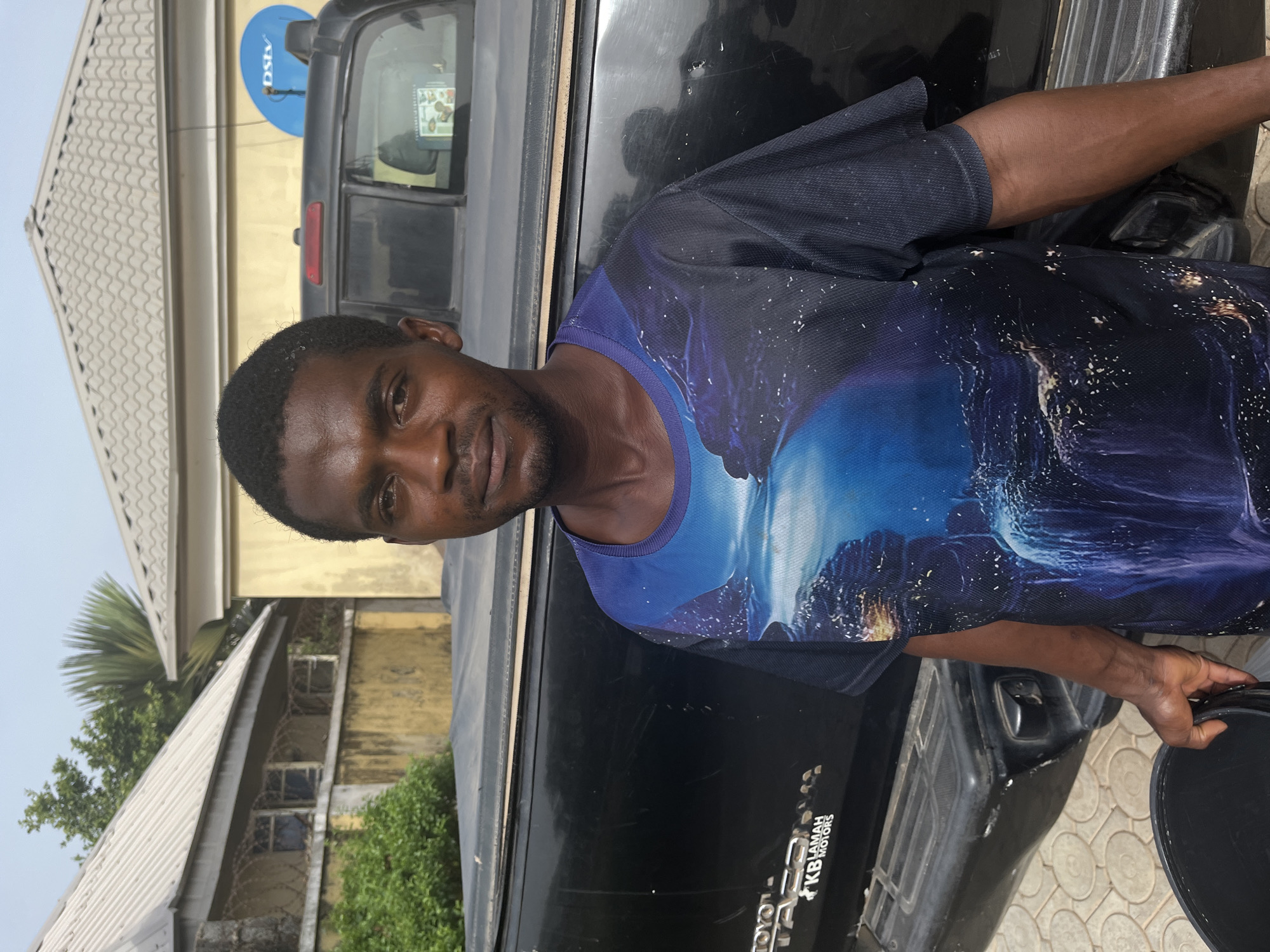 A man stands beside a black Toyota Hilux pickup truck in what appears to be a residential area. The man has short black hair and a trimmed beard. He wears a dark blue T-shirt with a printed design of a celestial landscape. He is holding a dark object, possibly a container. The truck has "KB Lamah Motors" on the lower right corner. Behind the truck are buildings with corrugated metal roofs, and one building has a satellite dish. The location is most likely in a residential neighborhood in Abuja, Nigeria, given the architectural style and presence of the car registration.