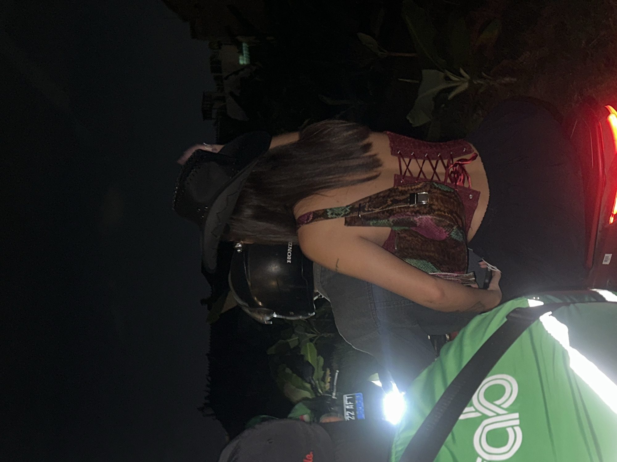 The image shows a night scene on a street in North Kuta, Indonesia. A person wearing a black cowboy hat and a red corset top is sitting on the back of a motorbike, holding a patterned handbag. The motorbike has a license plate that reads "22 AFT". Another person in a black helmet is riding the motorbike. To the left, part of a person wearing a green Grab jacket is visible. The environment appears to be tropical, with lush vegetation visible in the background. The street is illuminated by the motorbike's headlights and taillight, as well as some ambient light from nearby buildings.