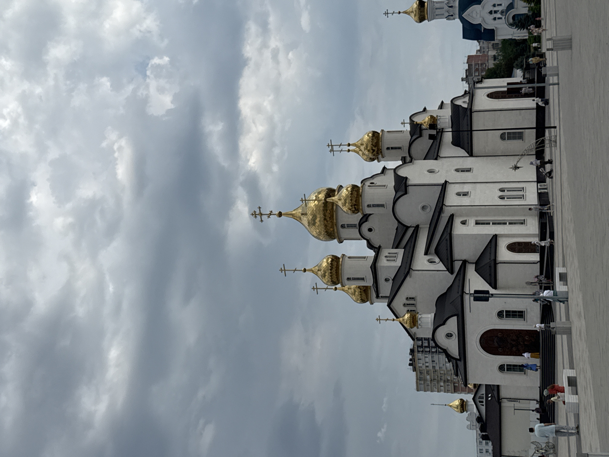 The image shows the Resurrection Cathedral in Anapa, Russia, under a cloudy sky. The cathedral is a large, white building with multiple gold-domed roofs, each topped with a cross. Several smaller golden domes are visible on the main structure, along with a taller, separate bell tower with a blue roof in the background. The cathedral is situated in a paved public square, where a few people are seen walking, sitting on benches, and standing. Some of these individuals are dressed in casual clothing, and a few are children. In the background, beyond the cathedral and square, are modern apartment buildings and trees. The overall scene depicts a moment of relative quiet in an urban setting, with the prominent religious structure as the focal point.
