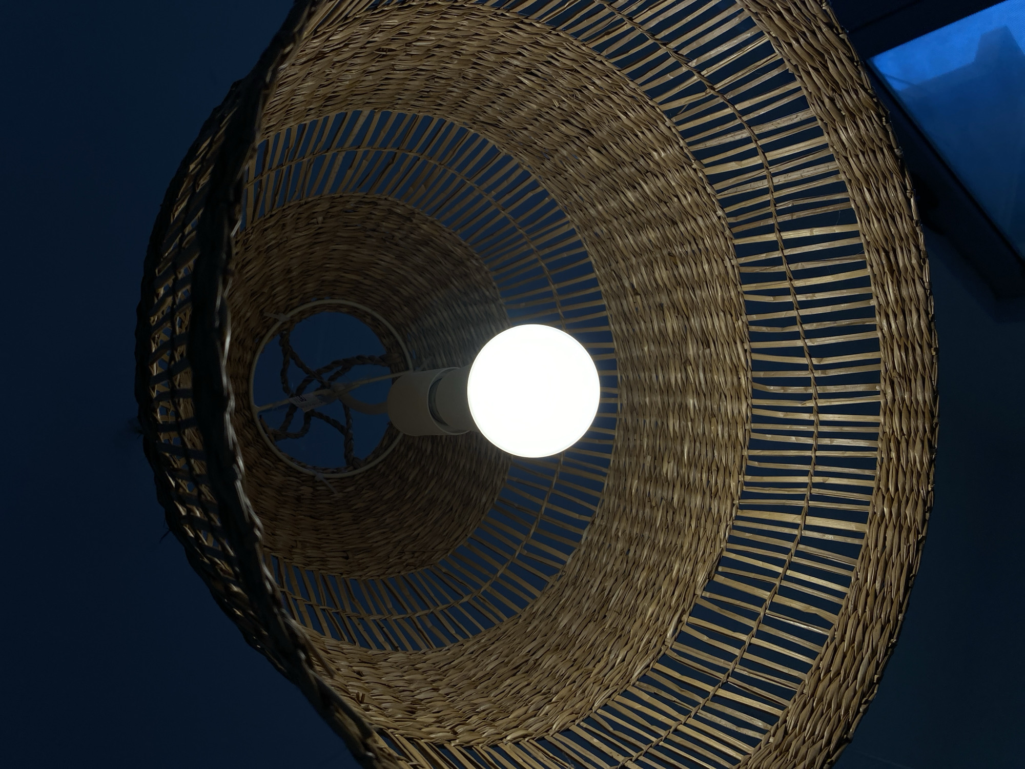 An illuminated light bulb is prominently featured in the center of the image, hanging from within a large, woven wicker lampshade. The light bulb is spherical, emitting a bright white light, and is attached to a white fixture. The lampshade is crafted from natural, light brown wicker material, intricately woven in concentric circles that create a spiral pattern when viewed from below. The weaving technique leaves small horizontal gaps between the rows, allowing glimpses of the dark background.
The perspective is looking upwards into the lampshade, creating a sense of depth as the wicker recedes into the background. The background itself is a deep, dark blue, suggesting either the night sky or a very dimly lit room. In the upper right corner of the original image, a slightly brighter blue area is visible, possibly a window or another light source reflecting on a surface.
No people or specific activities are visible. The scene is static, focusing on the decorative light fixture. The overall aesthetic of the woven wicker suggests a rustic, natural, or possibly coastal style, fitting for a location like Budva, Montenegro. There is no visible text in the image. The time of day appears to be night or late evening, given the prominence of the artificial light source against the dark surroundings.