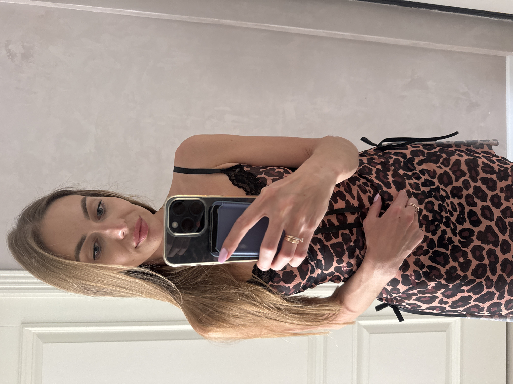 The image shows a woman with long blonde hair taking a mirror selfie. She is wearing a leopard-print slip dress with black lace trim and spaghetti straps. Her left hand is resting on her stomach, and her right hand is holding a smartphone. She is wearing two rings on her left hand. The background appears to be an indoor setting with a white door on the left and a textured wall on the right, possibly in a hotel room or apartment. There is no visual information to suggest the location is Sochi, Russia.