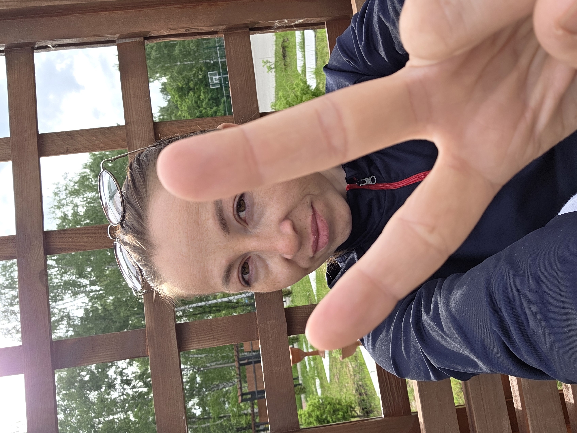 A selfie of a woman is taken from a low angle in an outdoor setting. The woman's face is visible between her fingers, as she makes a gesture toward the camera. She has brown hair and brown eyes, and sunglasses are perched on her head. She is wearing a navy blue jacket with a red zipper. The background shows a wooden structure, likely a gazebo, with greenery and trees visible through the gaps. In the distance, a basketball court can be seen. The exact location within the city of Unknown, Unknown, is indeterminable from the image.
