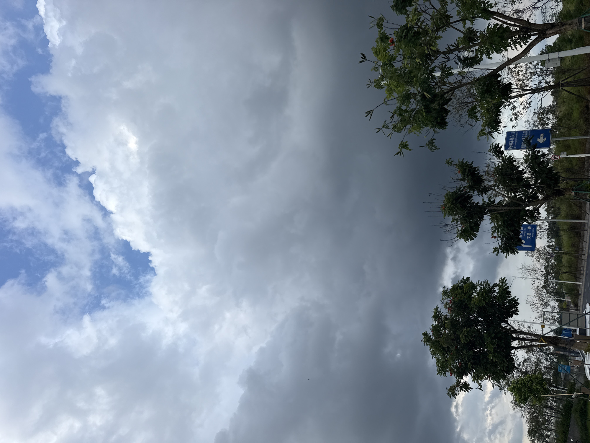 The image shows a cloudy sky over a road in Huizhou, China. In the foreground, there are several trees. On the right side of the road, there are blue traffic signs. The sky is dramatic, with dark storm clouds gathering in the lower half and lighter clouds and patches of blue sky in the upper half. There are no people visible in the image. The overall impression is one of impending weather change.
