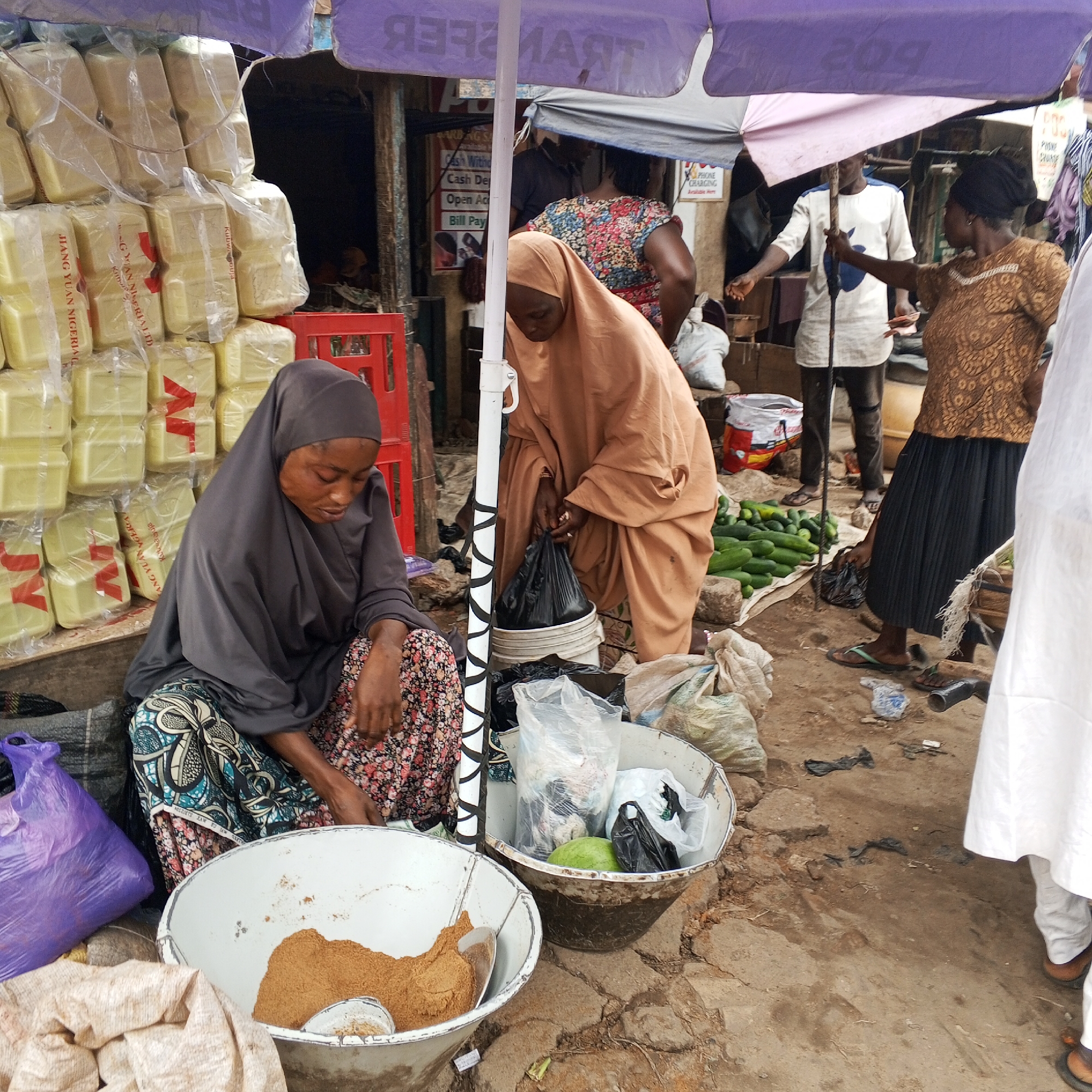 An outdoor market scene in Maiburiji, Nigeria, depicts several individuals engaged in commercial activities under the partial shade of large umbrellas. In the foreground, a woman wearing a dark grey hijab and patterned skirt sits on the ground, focused on a large white basin filled with a light brown powdered substance, possibly a spice or foodstuff. Adjacent to her, another woman in a light brown hijab stands, tending to goods in a bucket, holding a black plastic bag.
Various market items are visible: tall stacks of pale yellow rectangular blocks, individually wrapped in plastic, line the background, some bearing the red text "JIANG YUAN NIGERIA LTD." Green cucumbers are piled on a mat on the ground. Other containers hold plastic-wrapped goods, including what appears to be a green fruit or vegetable. A purple umbrella, reversed in some sections, displays text like "TRANSF" (likely "TRANSFER") and "POS," indicating a financial service point. Another umbrella pole features a distinctive black and white zigzag pattern.
In the mid-ground, other people are seen: a man in a white and blue tunic stands near the umbrella pole, and a woman in a brown patterned top and black pleated skirt appears to be engaging in a transaction, holding small items in her hand. A person in white attire is partially visible on the far right. The ground is unpaved and uneven. Signs in the background offer services such as "Cash Withdraw," "Cash Deposit," "Open Account," "Bill Pay," and "PHONE CHARGING." The overall atmosphere is that of a busy, functional market.