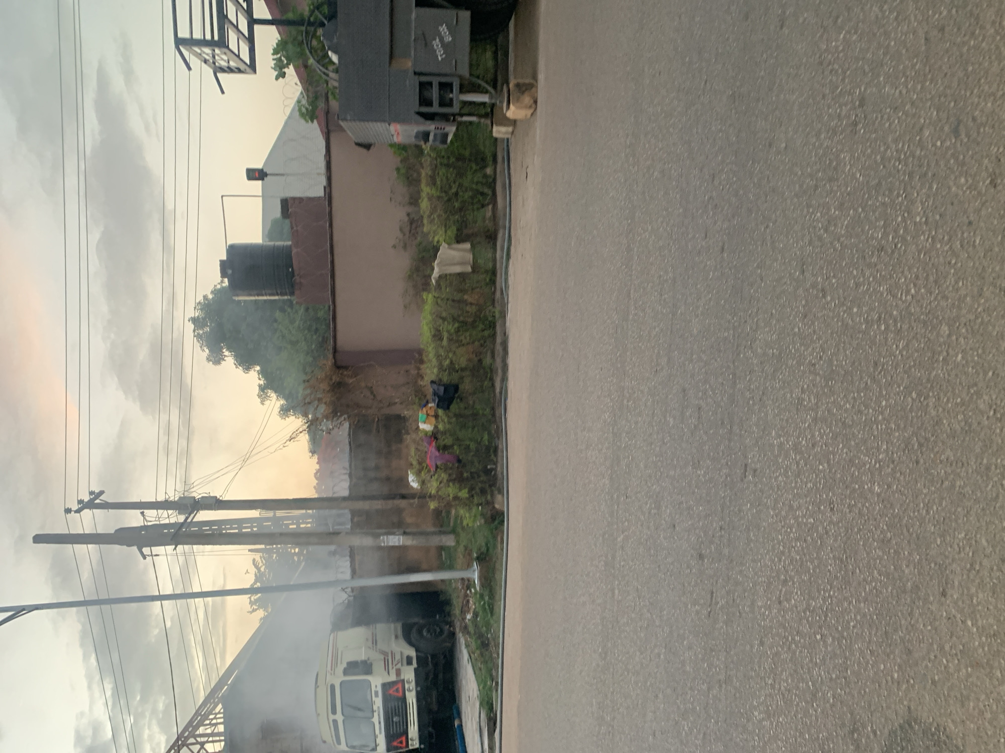 The image shows a street scene, likely in a residential area. There is a road or street in the foreground. On the left side of the image, there is a vehicle partially visible amidst smoke or vapor. Utility poles with wires are also present. Behind the vehicle and poles are houses or buildings, some with water tanks on the roof. Vegetation is visible. The sky appears overcast. It is not possible to confirm if this image is taken in Kabong, Nigeria.