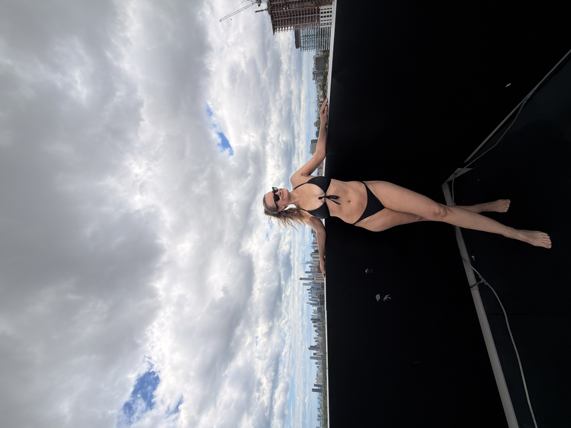 A woman in a black bikini stands on a rooftop in Toronto, Canada, leaning on a black barrier. She is wearing sunglasses and her hair is pulled back in a ponytail. Behind her, the Toronto skyline is visible, with numerous buildings and a construction crane. The sky is cloudy.