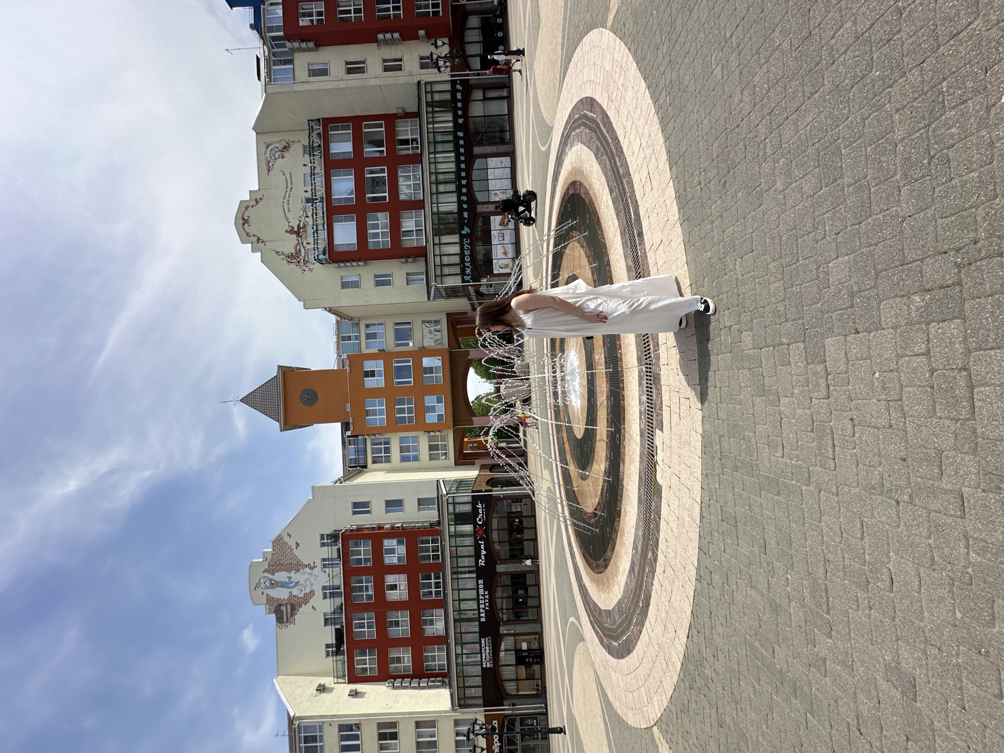 A young woman in a white dress stands near a circular fountain in a city square. The square is paved with grey cobblestones and features a fountain with spraying water in the center. Surrounding the fountain is a circular mosaic pattern. In the background, there are colorful buildings with a clock tower. The sky is blue with some clouds. A person on a scooter is visible in the background, and other people are walking in the distance. The image was taken in Krasnodar, Russia, specifically in the Sunny Gallery shopping center's adjacent area.