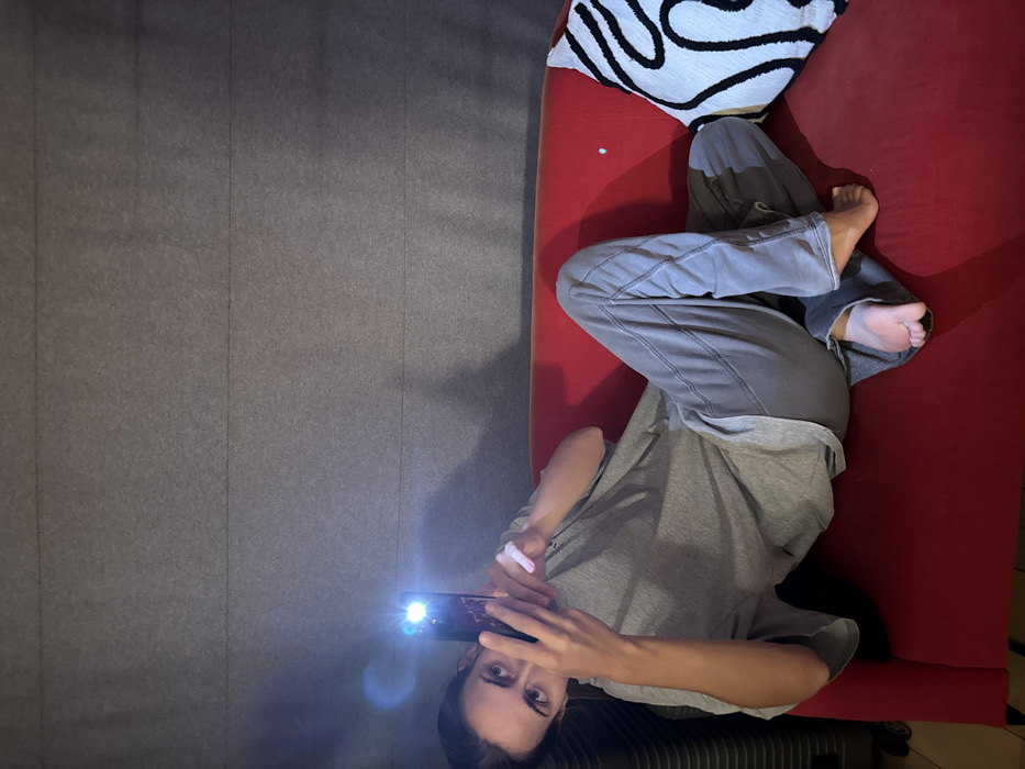 The image displays a single individual, appearing female, reclining on a red, upholstered sofa in an indoor environment. The person wears a grey short-sleeved t-shirt and grey sweatpants, with their right bare foot visible on the sofa. Both hands are holding a dark-colored smartphone in front of their face, pointed directly towards the camera. The phone's integrated flashlight is active, emitting a bright white light that creates a prominent lens flare. The individual's gaze is directed forward, towards the camera. The background consists of a dark grey wall with horizontal paneling, possibly acoustic or decorative. A white cushion with a black abstract or curvilinear pattern is positioned on the right side of the sofa. A dark, vertically ribbed object, potentially luggage or equipment, is partially visible on the far left edge of the frame. Shadows from the individual and the phone's light source are visible on the wall. The specific location within the city of Warsaw, Poland, cannot be determined from the visual information provided.