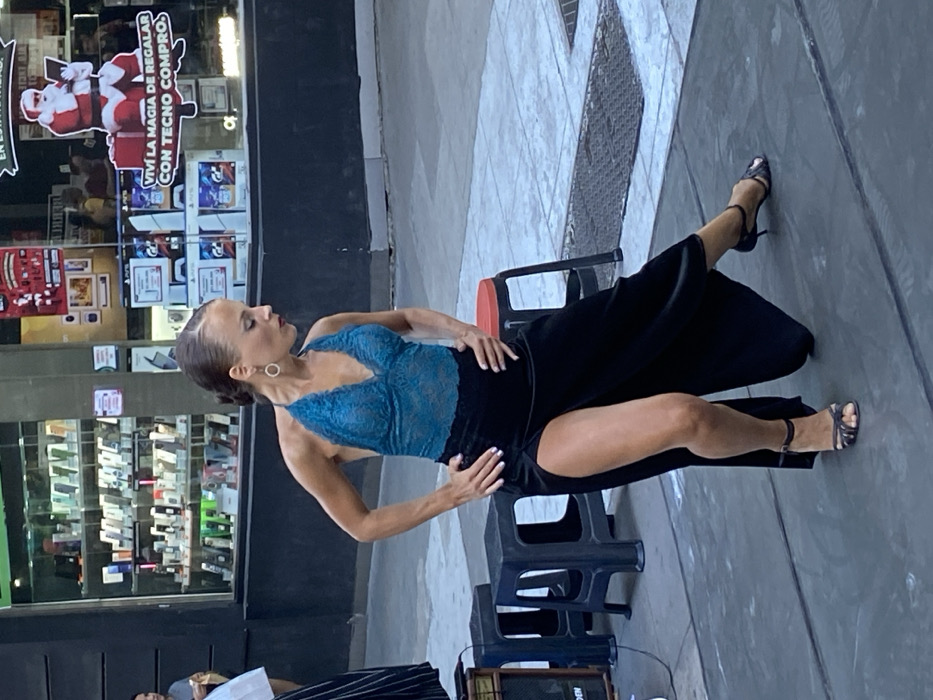 A woman in a blue lace top and black pants with a thigh-high slit poses outdoors. She wears strappy heels and earrings. A small black stool is near her. The background features a store window filled with merchandise and a Santa Claus display. The scene appears to be a street performance. The image has a slightly tilted perspective.
