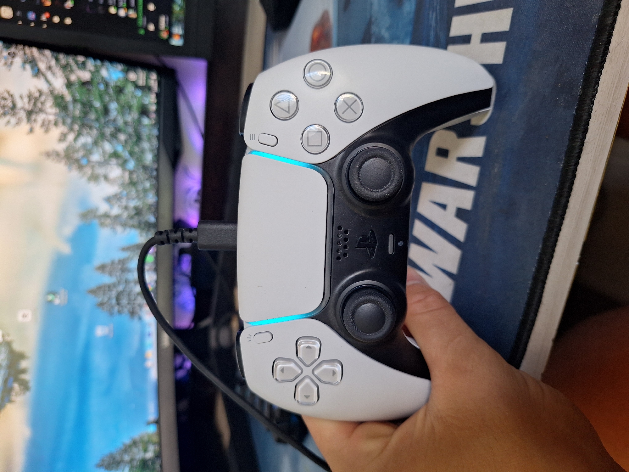 An off-center, eye-level shot shows a white video game controller being held in a person's left hand. The controller is a PlayStation 5 DualSense controller, with a USB-C cable plugged into the top. The controller has a light blue glow emanating from the strip above the controller buttons. In the background, a computer monitor displays a nature scene with trees and a blue sky. The controller rests on a dark blue mousepad with white lettering that appears to spell out "WAR". The lighting is natural, casting soft shadows. The scene is indoors, likely in a home or office environment. There is no indication of the city of Varna, Bulgaria in the image.