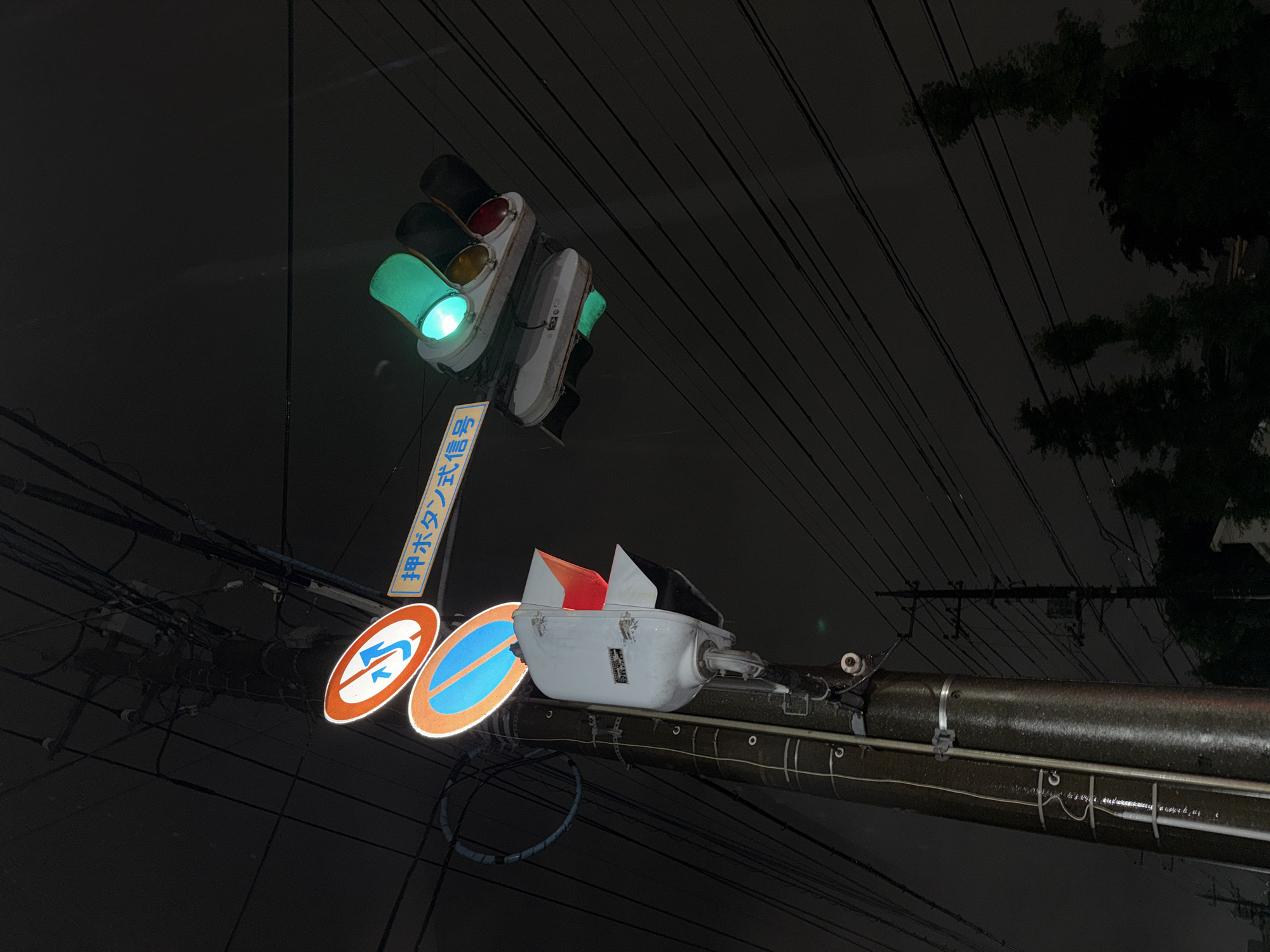 This image captures a nighttime scene in Yachiyo, Japan, likely on a street or intersection. The primary subjects are traffic signals and road signs, set against a dark, overcast sky with visible power lines crisscrossing the frame. One traffic light, angled downwards, prominently displays a bright green light. Another traffic signal is visible higher up, with its red and yellow lights illuminated. Below the traffic lights, two circular road signs are mounted on a pole. The sign on the left depicts a red circle with a blue arrow indicating a left turn, crossed by a diagonal red line, signifying "no left turn." The sign on the right is predominantly blue with a white horizontal bar, likely indicating a parking restriction. A blue and white sign with Japanese text is also visible, reading "押ボタン信号" which translates to "push button signal." The overall mood is somewhat somber, possibly enhanced by the rain, as the traffic signal housing and power lines appear wet. The presence of the traffic signals and road signs clearly indicates an urban or suburban setting where traffic management is in place.