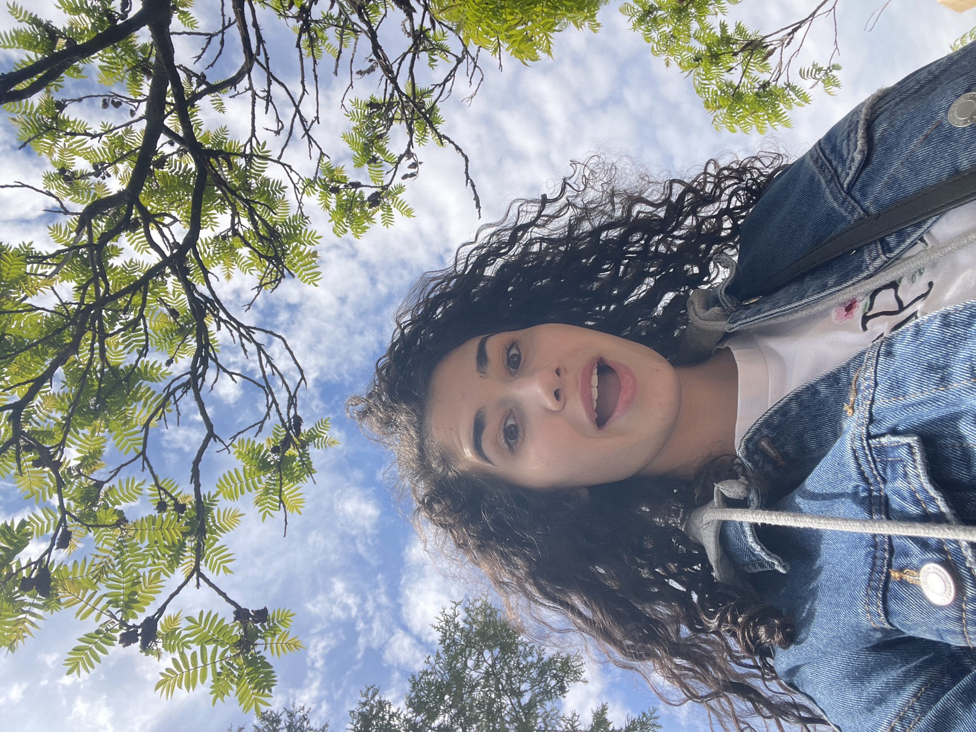 A low-angle, upward-facing photograph depicts a person, likely a young woman with dark curly hair, wearing a denim jacket over a white shirt. Her mouth is open in an expression of surprise or excitement. Above and around her are branches of trees with green foliage against a backdrop of a partly cloudy sky. Given the presence of deciduous trees, the photo likely was taken in a park or other green space. Determining the specific location in Sofia, Bulgaria from the visual information provided is impossible.