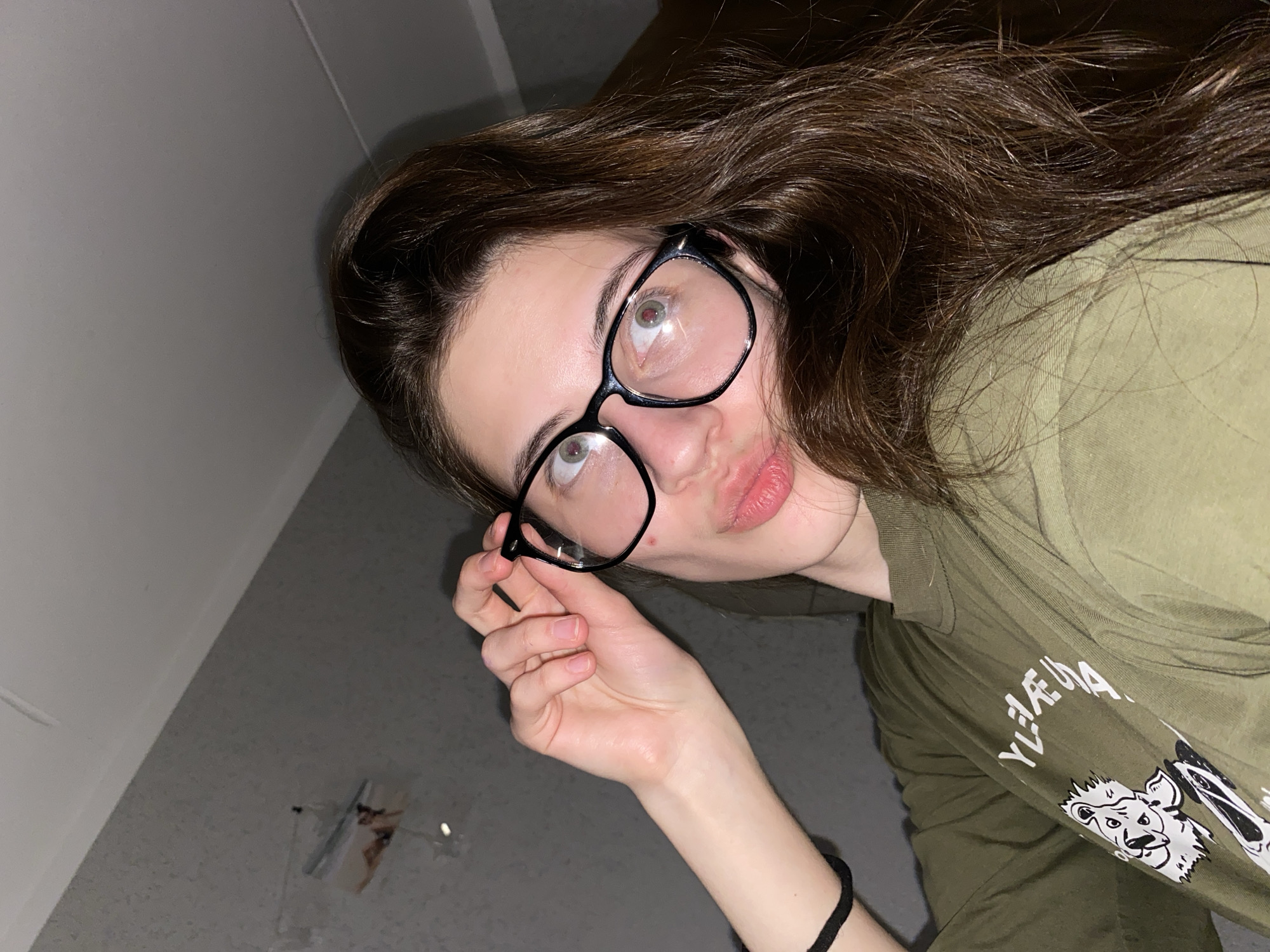 A woman with long brown hair is wearing black-rimmed glasses and an olive green shirt with a cartoon graphic. She is holding the glasses with her right hand. She has a black bracelet on her right wrist. The background consists of a gray wall with a white wall edge and what appears to be a piece of tape. The lighting is dim. There is no clear evidence to place this image specifically in Votkinsk, Russia.
