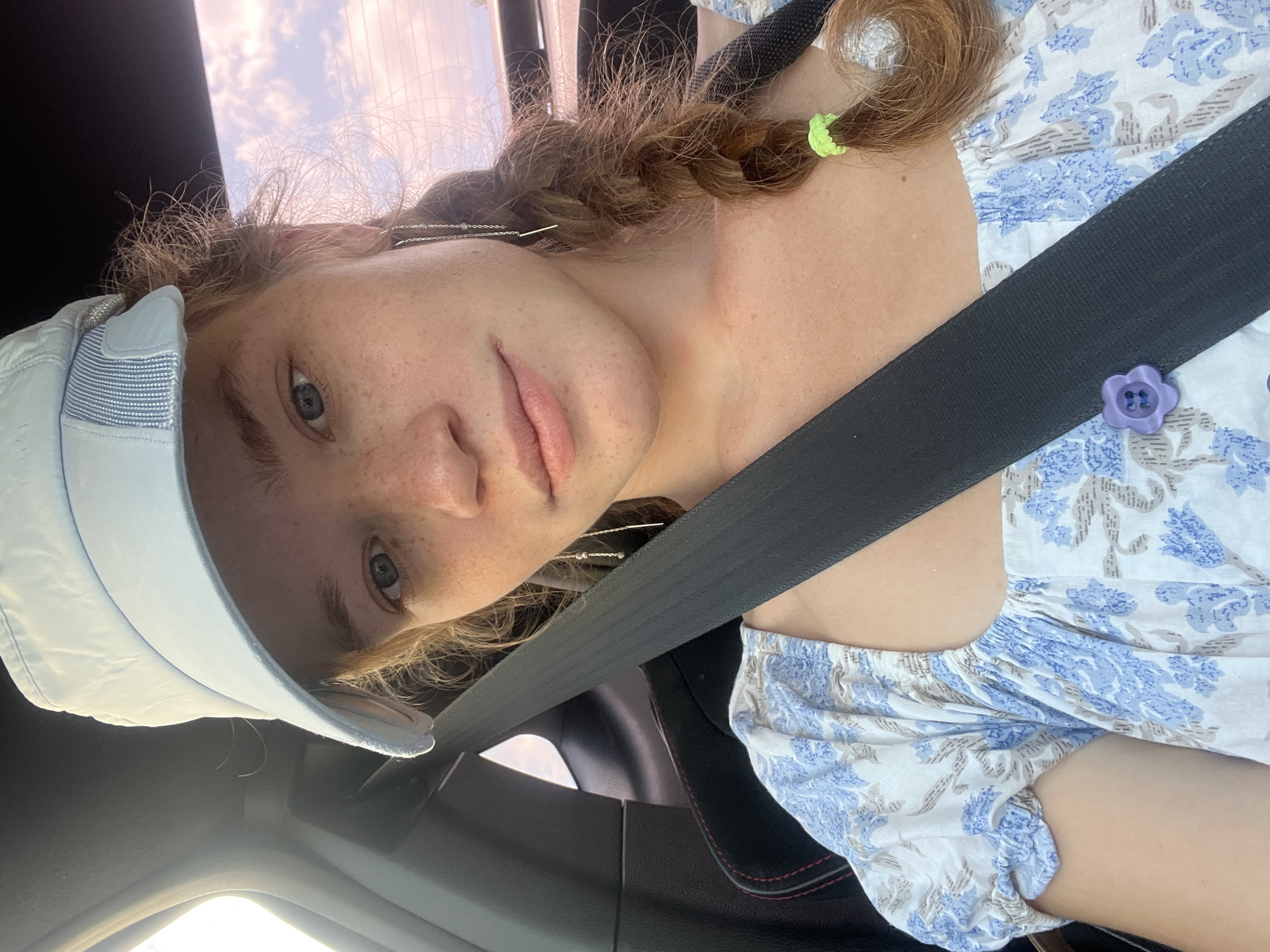 A young woman with reddish-brown hair braided and a light blue cap sits in the passenger seat of a car, wearing a blue and white floral print top and a seatbelt. She has freckles on her face and is wearing long, dangling earrings. A light purple flower-shaped button is visible on her dress, partially obscured by the seatbelt. The background shows a blurred view of the sky with clouds, suggesting the car is in motion. The interior of the car, including the black seat and seatbelt, is also visible. The scene appears to be outdoors, possibly on a road.