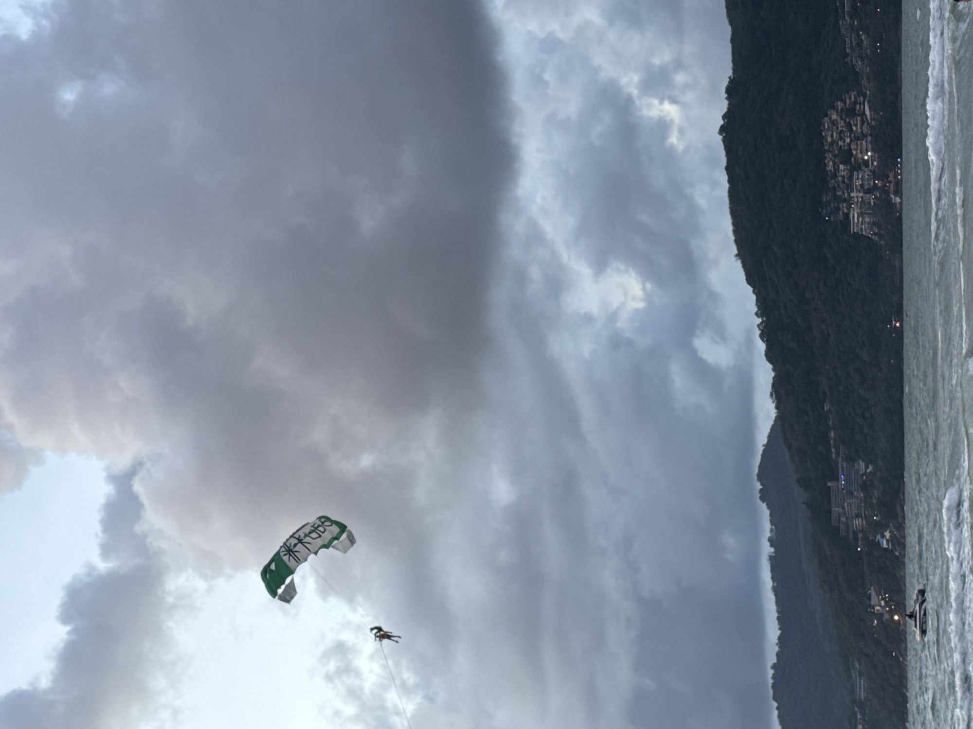 The image depicts a parasailer in mid-air above the ocean at Patong Beach, Thailand. The sky is overcast with dramatic, dark clouds. The parasail, a large green and white parachute, is clearly visible with some text on it. A single person is suspended beneath it, attached by lines. In the background, a lush, green, mountainous coastline is dotted with buildings, some of which are illuminated, suggesting a coastal town. The sea in the foreground is choppy with visible waves. A small boat with at least one person on board is visible on the water. The overall atmosphere is moody, likely due to the overcast weather conditions.