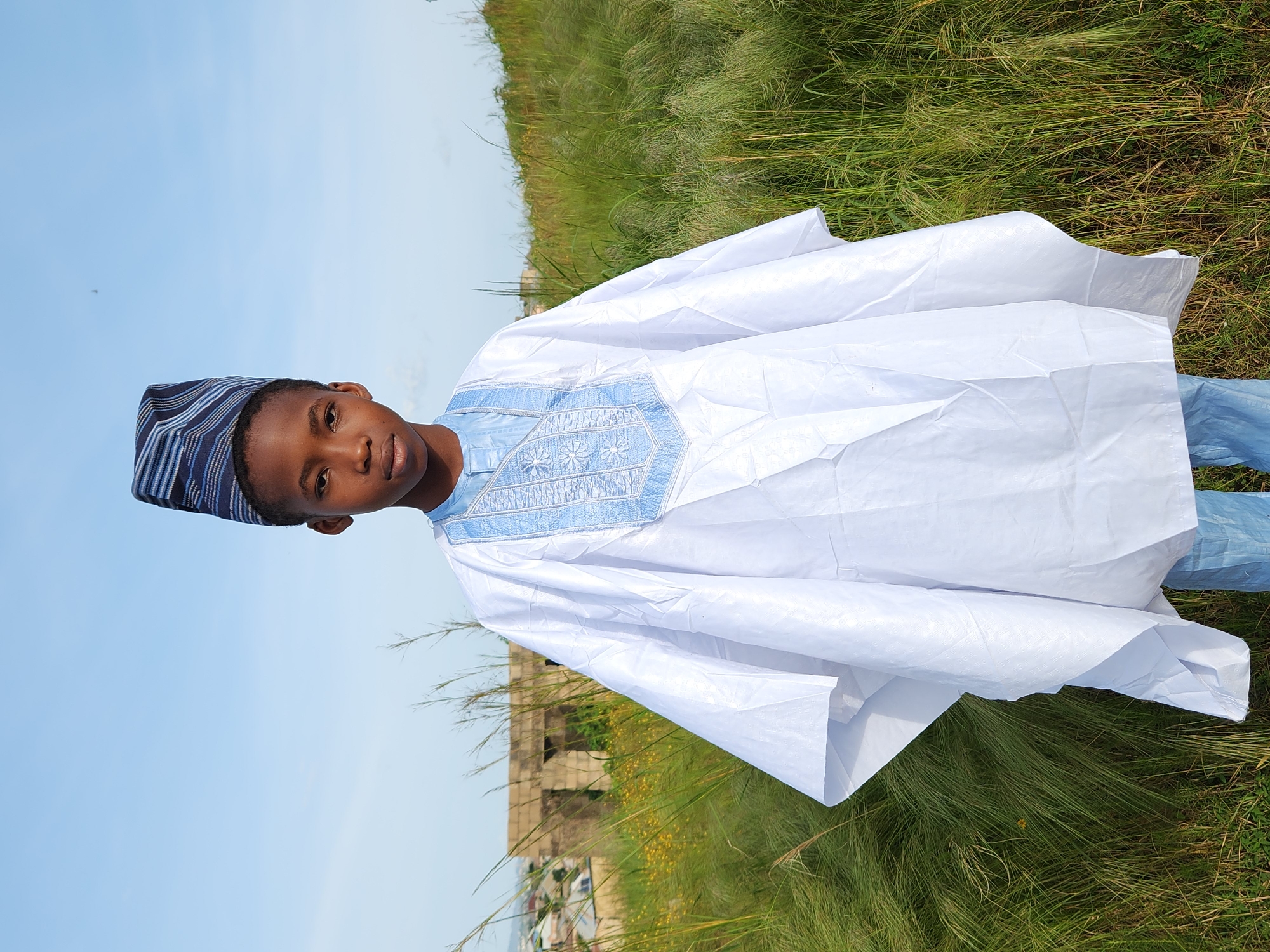 A young boy stands outdoors, facing the camera, in a grassy, hilly area. He is wearing a traditional white boubou with blue embroidery around the neckline, and a patterned blue and white cap. The boy has dark skin and is looking directly at the camera with a neutral expression. Behind him, a partially constructed stone building is visible on a hillside, with a town or city spread out in the background. The sky above is a clear, light blue. The overall impression is a portrait of a child in traditional attire in a semi-rural setting, possibly in or near Jos, Nigeria, given the architectural style and landscape.