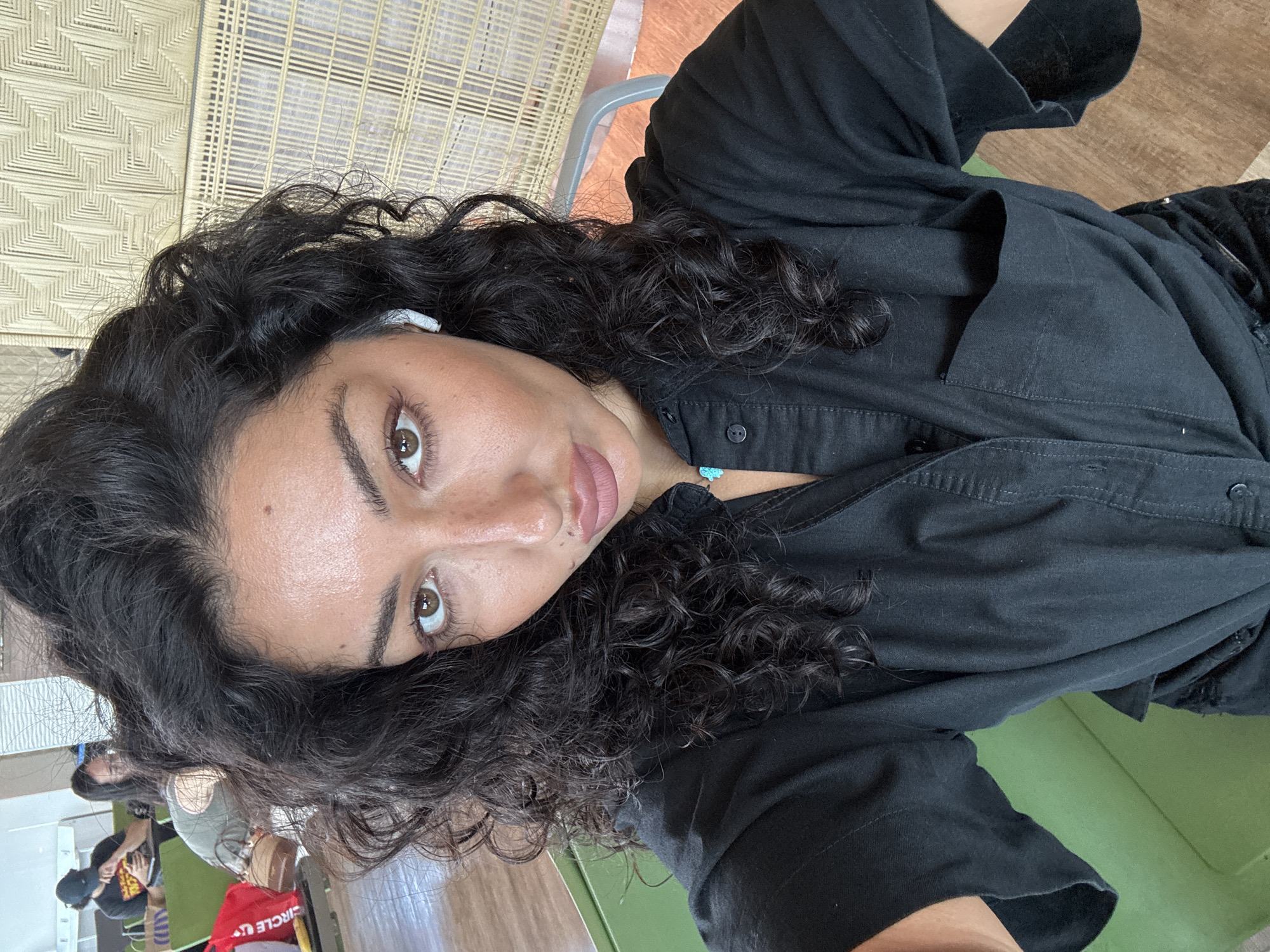 An image, captured with a tilted perspective, features a young woman taking a selfie, likely indoors. She has dark, voluminous curly hair that frames her face and shoulders. Her skin has a warm tone, and she looks directly at the camera with large, brown eyes. Her eyebrows are well-defined, and she has natural-looking makeup, with a touch of color on her lips.
She is wearing a dark, possibly black, collared button-up shirt. A small white earbud is visible in her right ear, and she wears a delicate light blue beaded necklace around her neck.
The background shows elements of an indoor setting. Behind her head and to the left, there's a light-colored woven surface with a distinct geometric pattern, resembling rattan furniture or a wall covering. Further into the background, there's a mix of a green surface (possibly a bench or floor) and a brown wooden floor. In the lower left corner, blurred figures of other people and a red bag with partially visible text "CIRCLE U" can be discerned, suggesting a public or communal space like a cafe or lounge. The lighting indicates it is daytime. The overall mood conveyed is calm and casual.