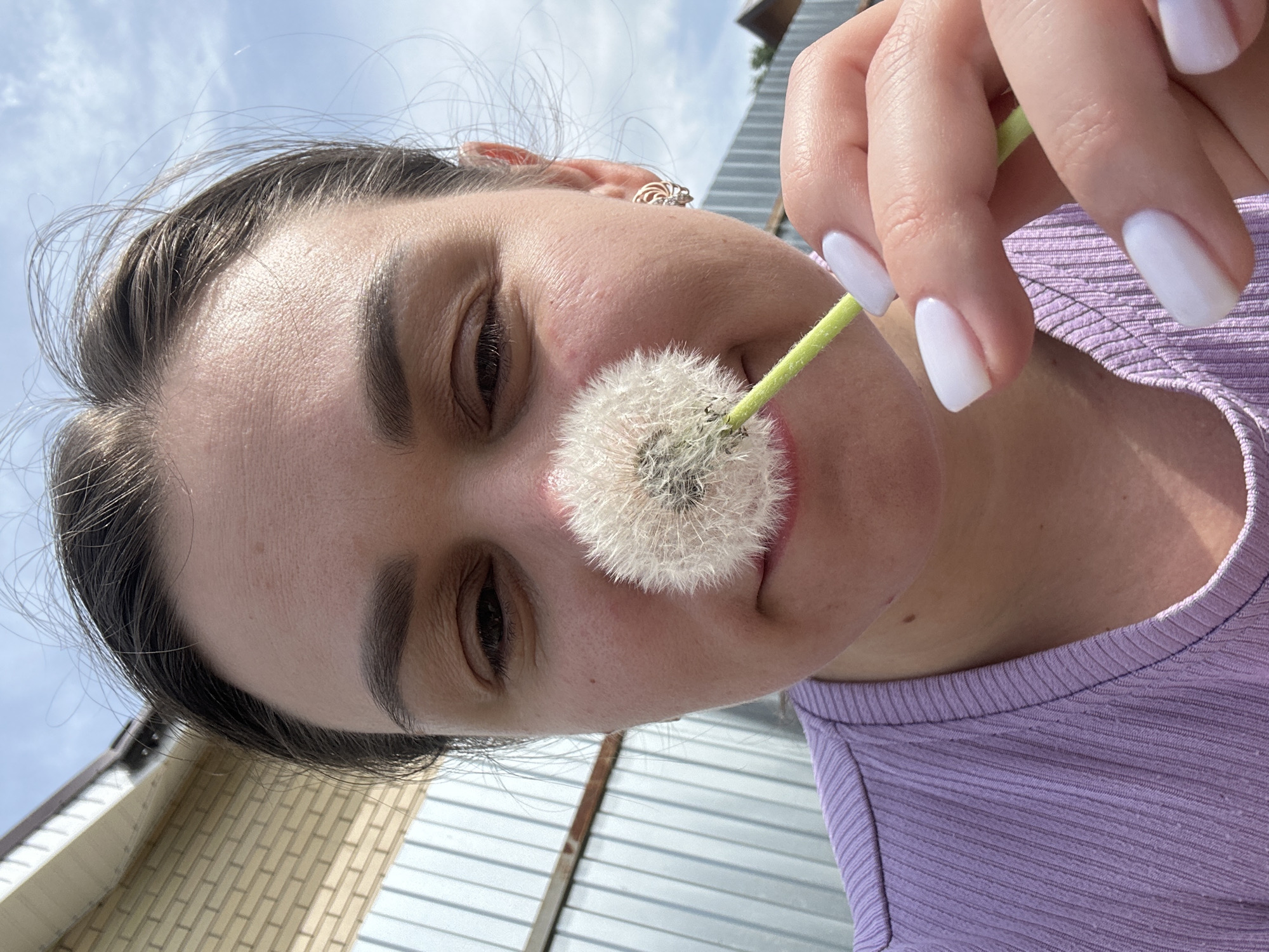 A woman is holding a dandelion in front of her face. The dandelion is white and fluffy. The woman has fair skin, brown eyes, and dark hair. She is wearing a light purple ribbed shirt. Her fingernails are painted white. She has a ring in her ear. The background is out of focus, but appears to include a blue sky with clouds, a brick building, and a metal fence. The location is Chaikovsky, Russia.