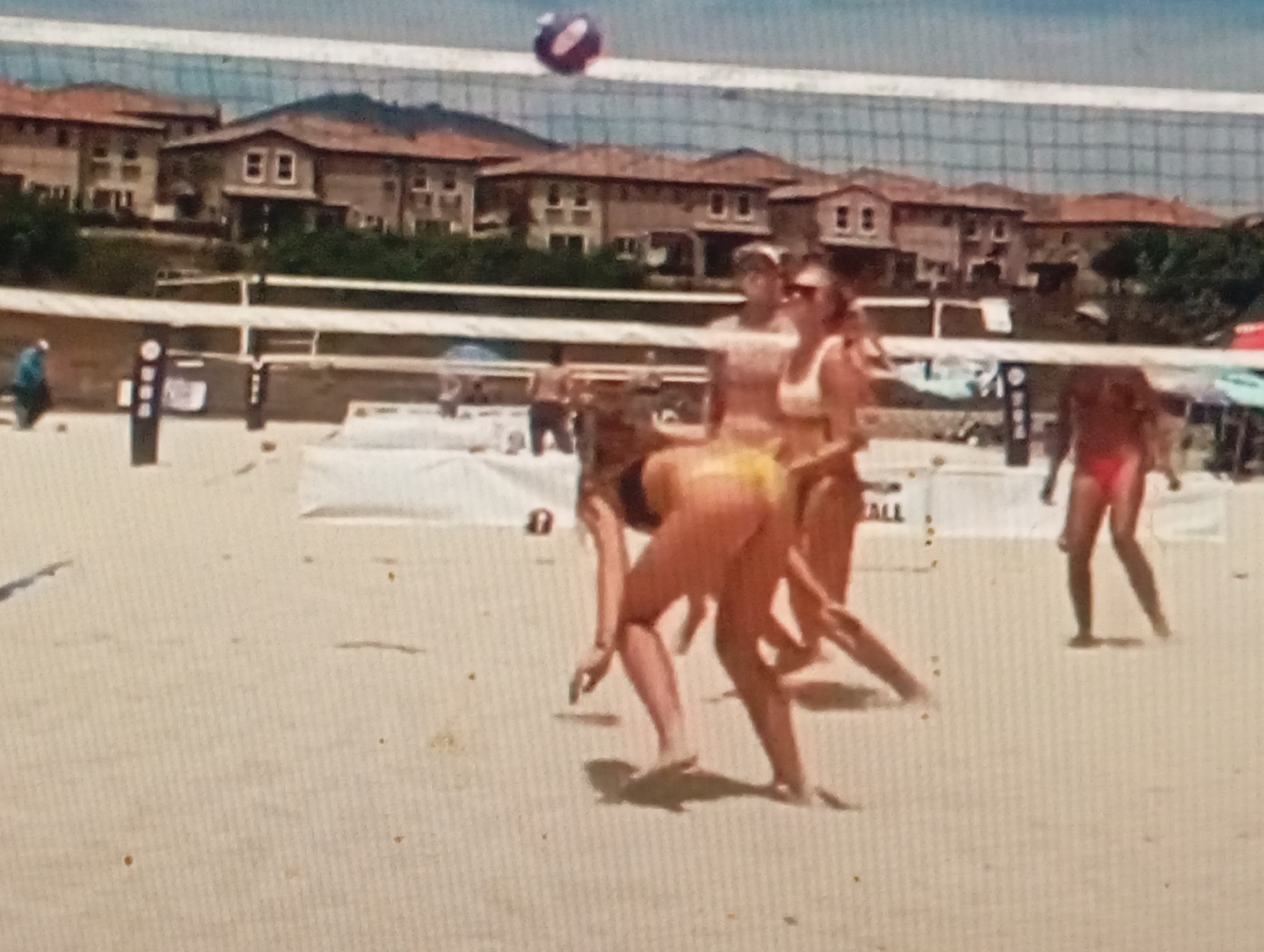 This image depicts a beach volleyball game in a sunlit outdoor setting. The main subjects are several individuals playing volleyball on a sandy court, with a net dividing the playing area. The players are dressed in swimwear, actively engaged in the game. In the background, a row of houses with red-tiled roofs is visible, suggesting a residential area near the beach. The sky appears bright and clear, indicating fair weather, likely during the daytime. The scene conveys a sense of recreation and sport. There is no visible text in the image.