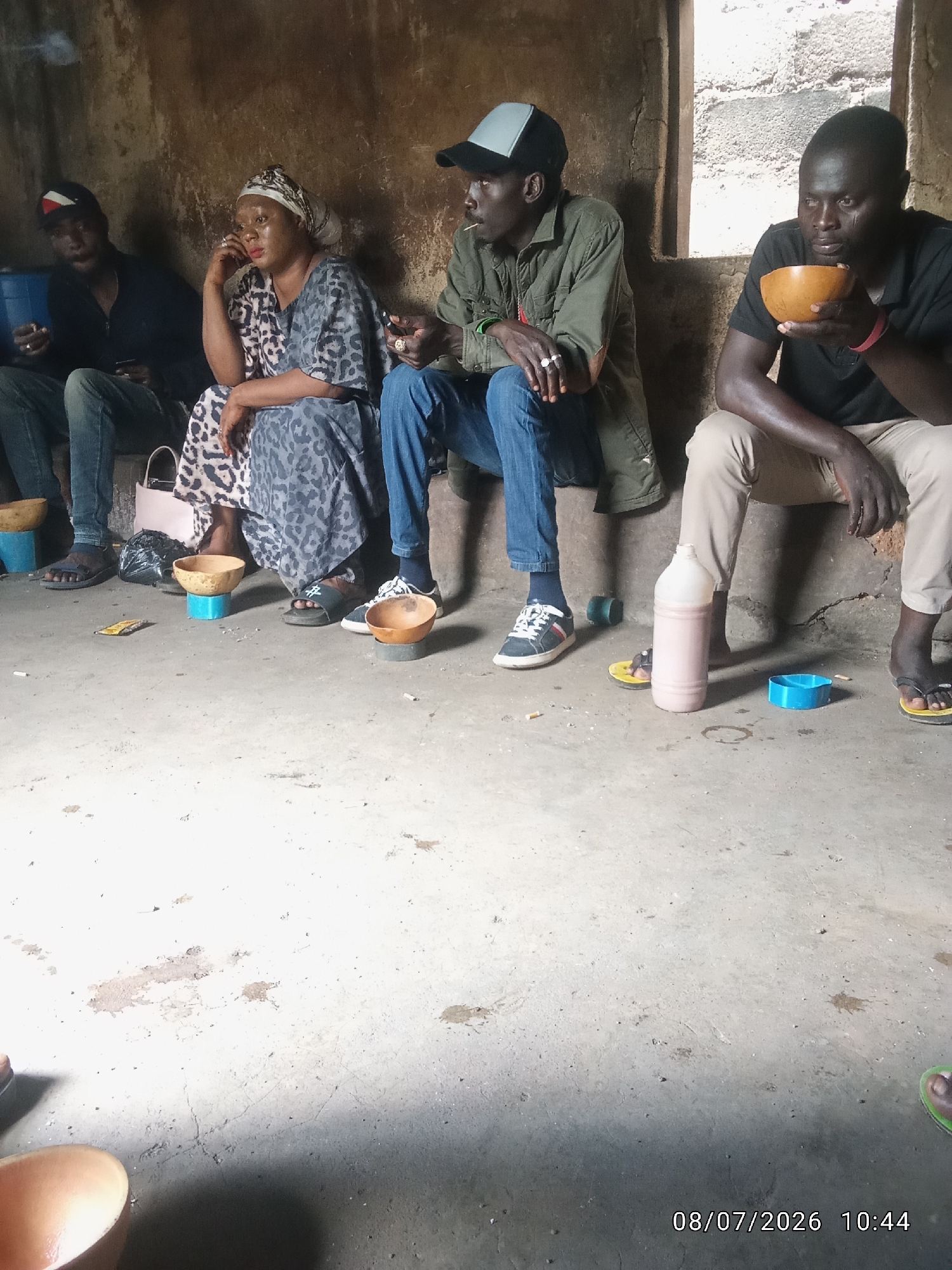 The image depicts a group of five individuals, four men and one woman, seated on the floor in what appears to be an interior space with a textured, earthy-toned wall and a concrete floor. The individuals are casually dressed and appear to be engaged in conversation or relaxation.
From left to right:
* The first person, a man, is partially visible, wearing a dark jacket and jeans. He is holding a phone and a cigarette.
* The second person, a woman, is seated with her legs crossed, wearing a patterned dress and a headscarf. She is touching her face with her right hand, which is adorned with rings.
* The third person, a man, is wearing a green jacket and jeans. He is holding a cigarette and has a ring on his left hand. He is also wearing a baseball cap.
* The fourth person, a man, is wearing a black polo shirt and light-colored trousers. He is holding a wooden bowl in his right hand and appears to be looking at it.
* The fifth person, a man, is wearing a black t-shirt and beige trousers, partially visible. He is wearing sandals.
Several objects are present on the floor:
* A blue plastic container is near the first person.
* Two wooden bowls, one supported by a blue base, are in front of the woman and the third person.
* A pink bottle is located in front of the fourth and fifth persons.
* A blue plastic container is near the fifth person.
* Several cigarette butts are scattered on the floor.
* A pink bag is visible behind the woman.
The environment is dimly lit, suggesting an indoor setting. The wall behind the individuals is rough and uneven. The floor is concrete and shows some stains and cracks. A date and time stamp at the bottom right of the image reads "08/07/2026 10:44". The location is noted as Unknown, Unknown.
