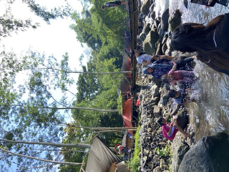 A daytime shot shows a group of people enjoying a rocky creek. Adults and children sit and splash in the water. A tent and a small building with a red tarp are visible on the left bank, along with trees and vegetation. A person with long hair stands near the water's edge, possibly taking photos with a phone. A person is walking on a structure in the distance.