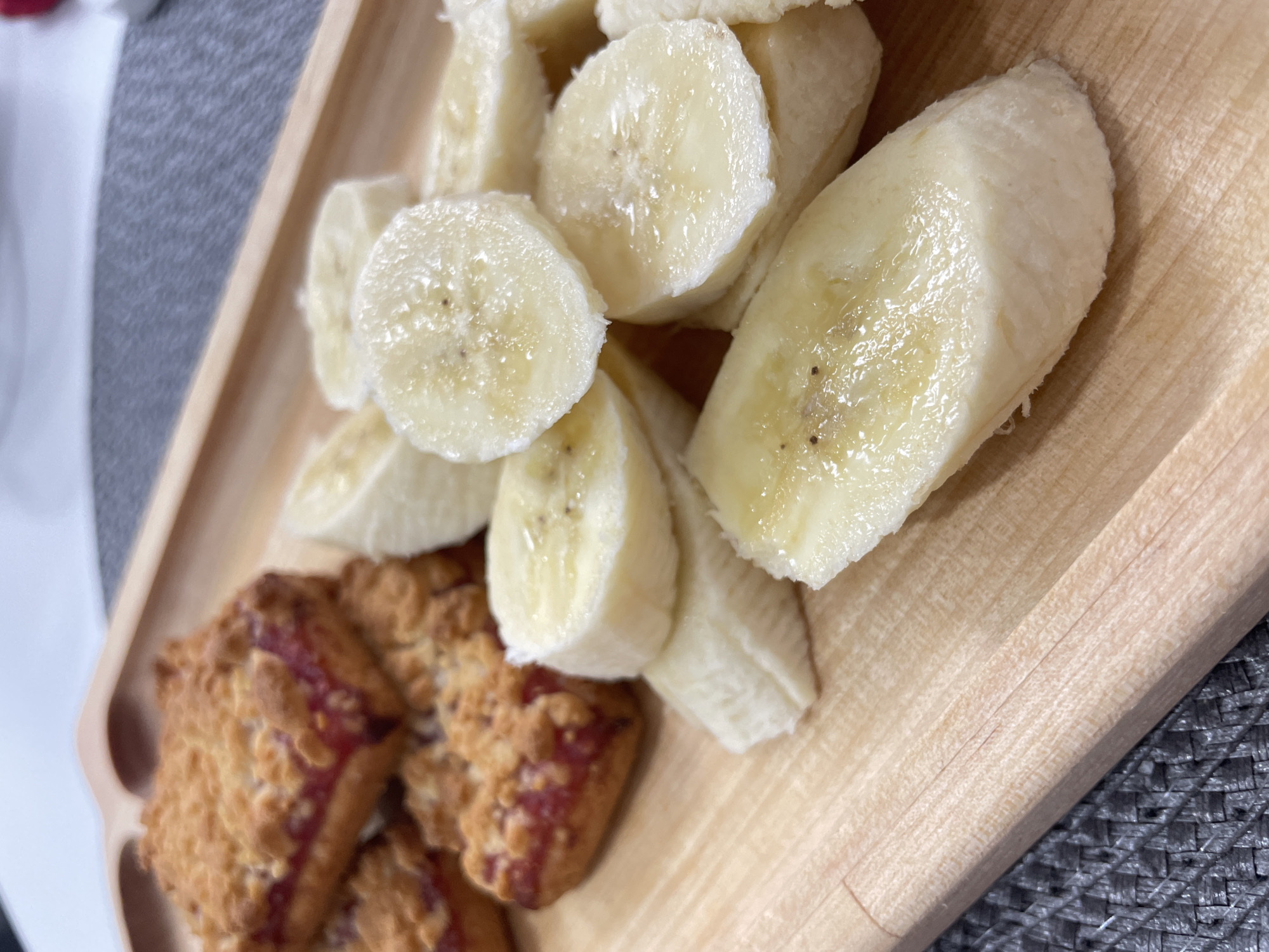 The image shows a close-up of a light wooden tray with sliced bananas and what appear to be square-shaped pastries or scones with a reddish filling, possibly jam. The items are placed on the tray in a seemingly informal arrangement. The tray is set on a gray woven surface. The absence of other details suggests this is a still life, likely photographed indoors. The location in Izhevsk, Russia, cannot be determined from the image alone. No people, actions, or interactions are visible.