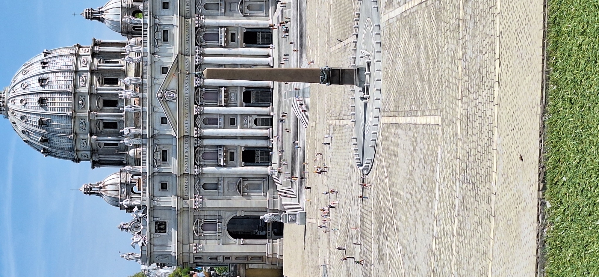 The image displays a miniature model of Saint Peter's Basilica and Square in Vatican City, Rome. The detailed model features the iconic dome, facade with columns and statues, and the expansive cobblestone square. A central obelisk stands in the square, surrounded by a circular arrangement of bollards. Numerous tiny figures, representing people, are scattered across the square and on the steps leading up to the basilica, suggesting a bustling scene. The foreground shows a strip of meticulously manicured green grass. The overall impression is of a well-crafted diorama designed to replicate the grandeur of the actual landmark. The location identified as Rimini, Italy, is incorrect for Saint Peter's Basilica.