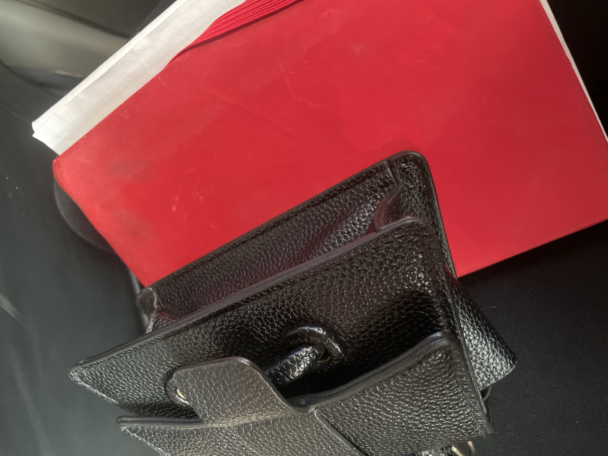 The image shows a close-up view of a red and white notebook overlaid by a textured black leather wallet. The wallet's flap and closure mechanism are visible. The items rest on a dark surface. The location of Kurunduma, Nigeria, is not evidenced within the frame.