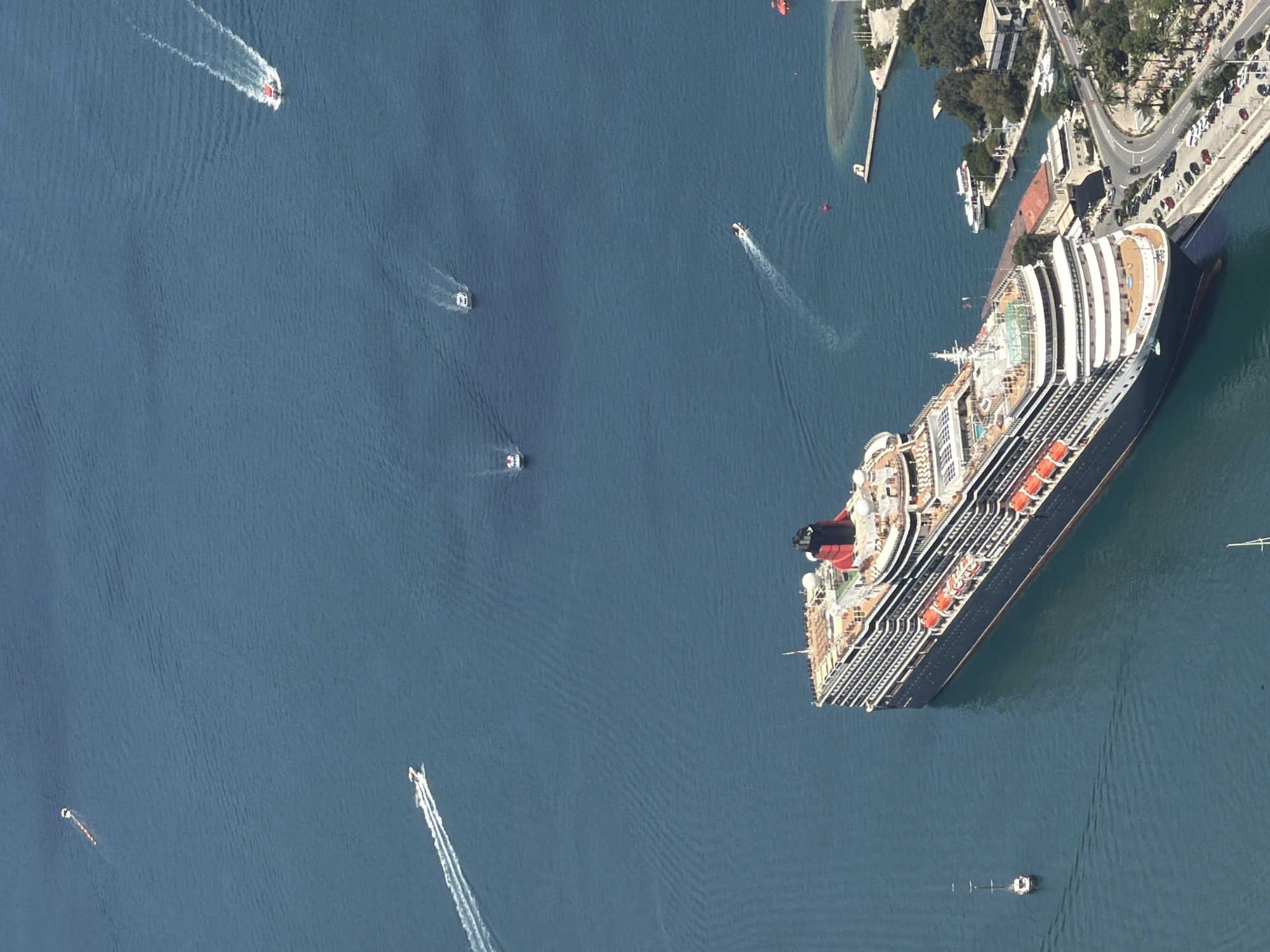 An aerial image provides a high-angle view of a large cruise ship near a coastal town, likely in Špiljari, Montenegro. The massive ship, featuring multiple decks, numerous lifeboats along its side, and a distinctive red and black funnel, occupies a significant portion of the lower right frame, appearing either docked or moored very close to land.
The calm, deep blue water around the cruise ship is bustling with activity from several smaller boats. These are mostly speedboats, each leaving a clear white wake, indicating movement. At least five such boats are visible; some are white, and one distinctively red/orange boat is seen in the upper left. A red and a yellow/orange buoy are also present in the water.
The coastline, visible in the upper right, features a developed area with a winding road, numerous parked cars, and various buildings, many with red-tiled roofs, nestled among green trees. A small, enclosed harbor or marina with additional small vessels is also part of the coastal landscape. The scene is bathed in bright daylight, suggesting clear weather. No visible text is discernible on any of the objects or structures within the image.