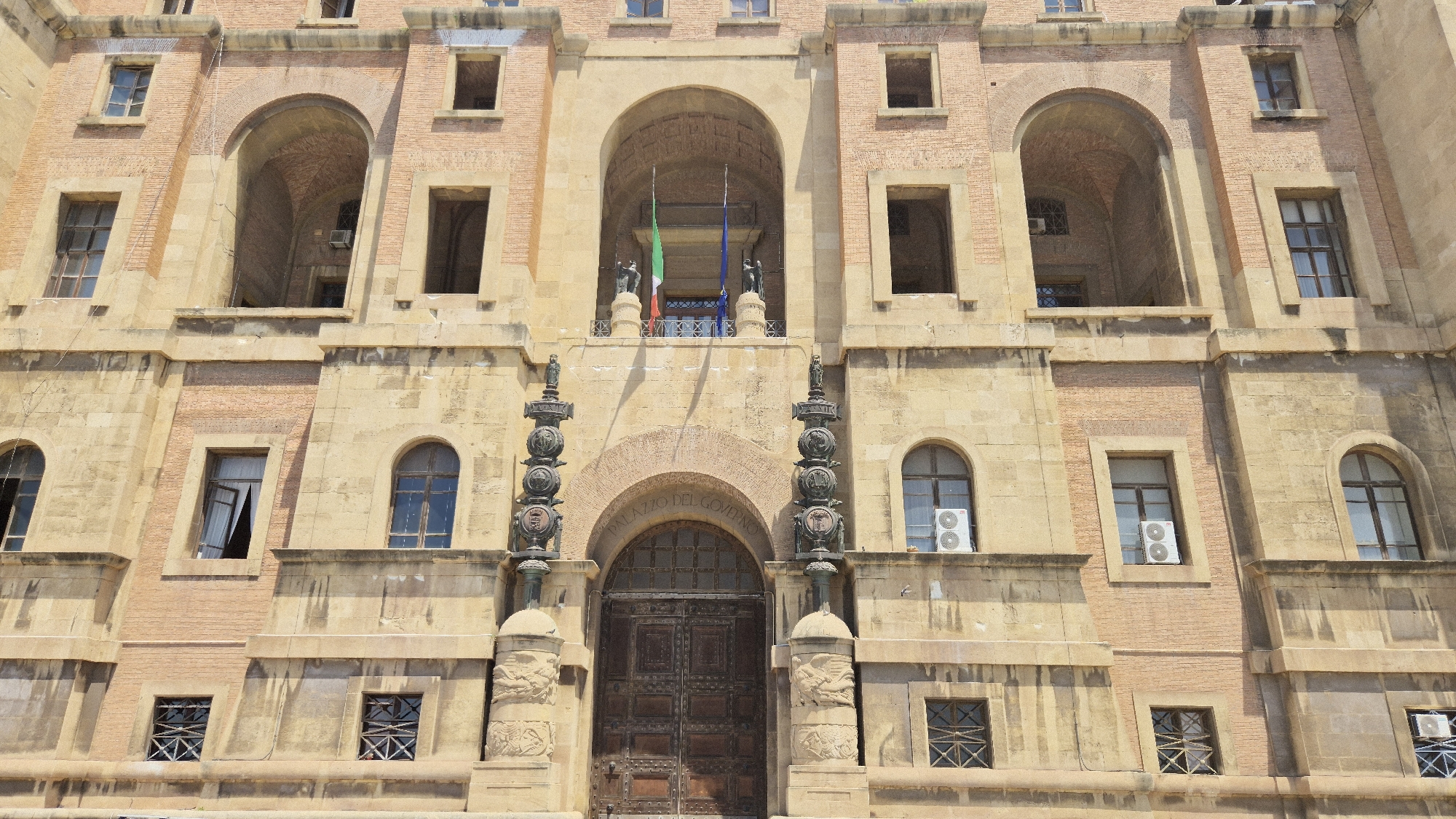 The image shows a frontal view of the Palazzo del Governo in Taranto, Italy. The building is made of light brown stone and red brick, with several arched windows and a large wooden door at the center. Two flags, one Italian and one European, are visible hanging from an arched balcony above the entrance. Two statues stand on either side of the entrance, flanking two large ornate lampposts. The overall scene depicts a historic public building in an urban setting.
