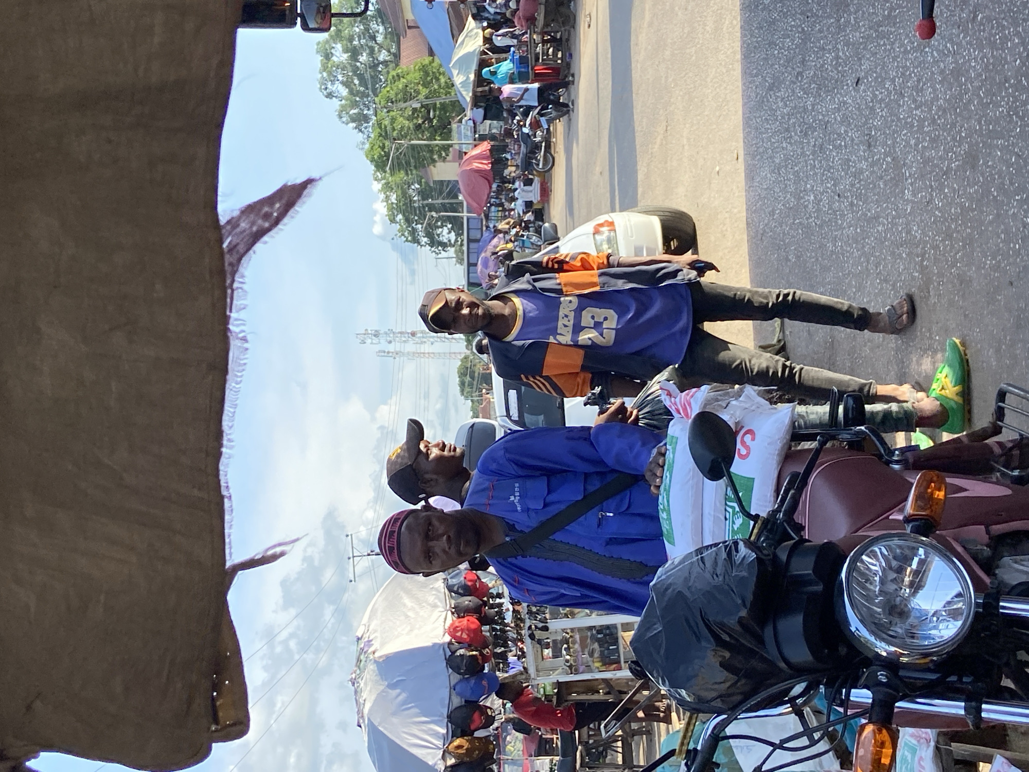 The image depicts a street scene in Mararaba, Nigeria. The view is from a vehicle, likely a car or a tricycle (keke), as indicated by the roof or canvas structure in the foreground. In the middle ground, two men are visible. One is wearing a blue Lakers jersey with the number 23, a black cap, black pants, and sandals. The other man is wearing a blue shirt, a skull cap, and has a bag slung across his shoulder. They appear to be interacting with a vendor or someone selling goods from a motorcycle. The motorcycle is in the foreground, loaded with bags. In the background, there's a crowded street market with various stalls covered by umbrellas and tarps. Cars and motorcycles are also visible amongst the crowd, indicating a busy commercial area. Power lines and utility poles are also visible. The lighting suggests a sunny day.