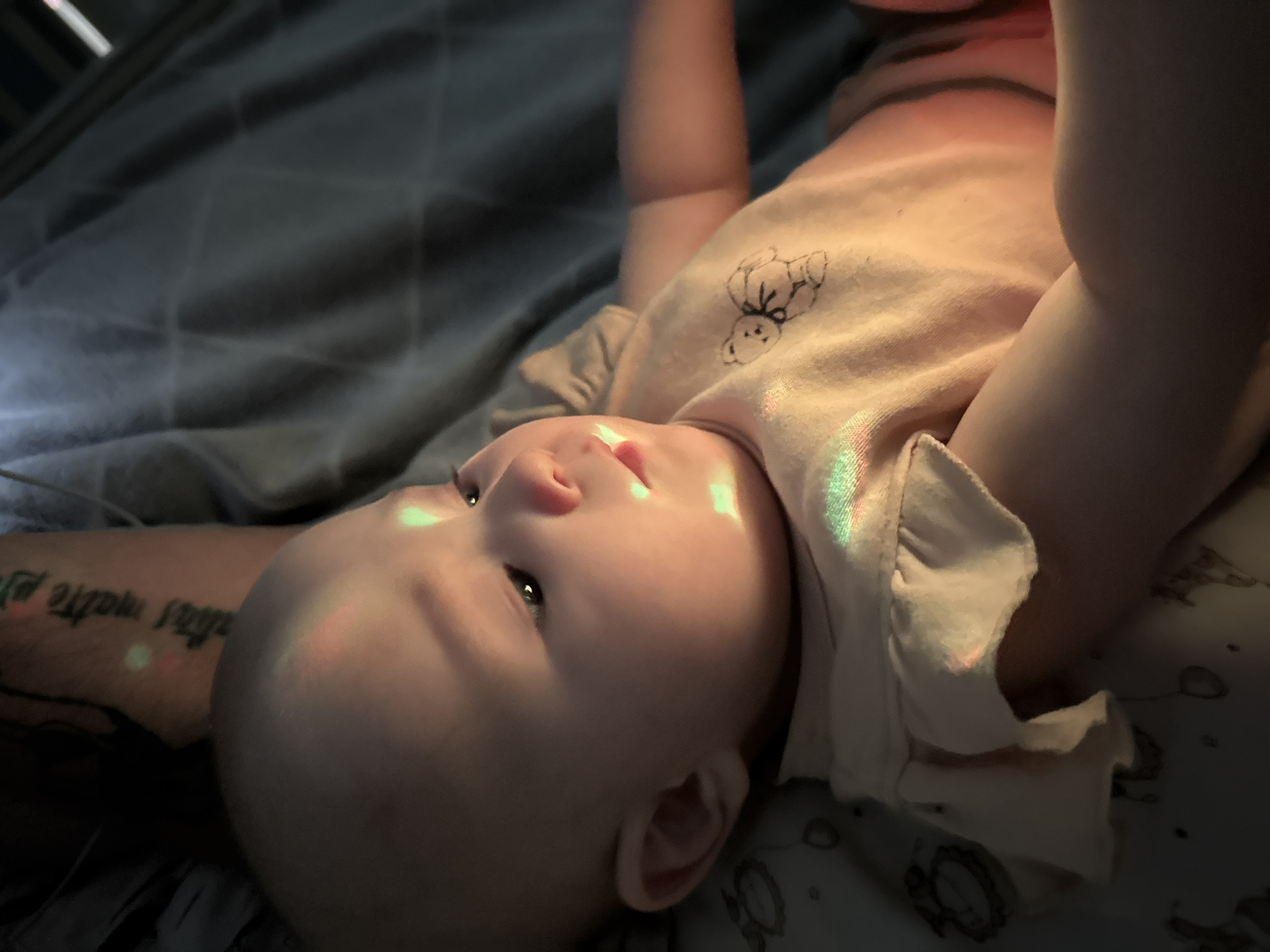 A close-up photograph captures a baby lying on its back, appearing to be on a bed. The baby is wearing a light-colored onesie adorned with a small bear illustration. Light patterns, possibly from a projector or prism, are cast on the baby's face and clothing. The background includes a gray blanket and an adult's tattooed arm. The location in Yakutsk, Russia cannot be determined from the visual information provided.
