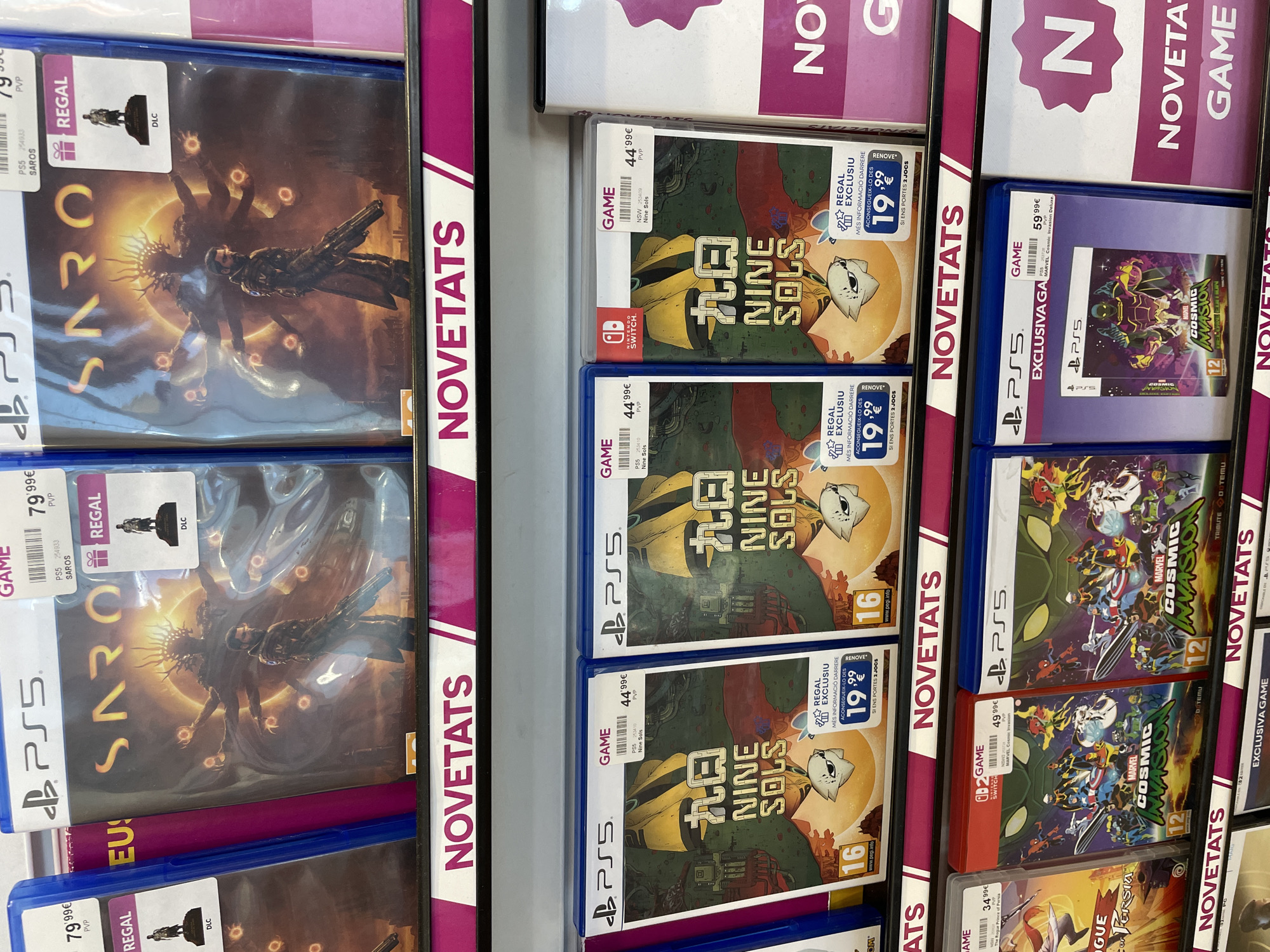 The image shows a close-up of a shelf in a store, likely located in Mataró, Spain, given the Catalan text on the shelf labels ("NOVETATS"). The shelf is stocked with video games for PlayStation 5 (PS5) and Nintendo Switch.
There are multiple copies of the PS5 game "SAROS" priced at 79.99€ each. There are also multiple copies of the games "Nine Sols" for both PS5 and Nintendo Switch, priced at 44.99€, with a promotional "RENOVE" sticker offering the game for 19.99€ if two old games are traded in. Additionally, there are copies of "MARVEL Cosmic Invasion Deluxe" for PS5, priced at 59.99€, and the standard version of "MARVEL Cosmic Invasion" for Nintendo Switch, priced at 49.99€. The games are arranged neatly on the shelf, with the "NOVETATS" label visible along the side.