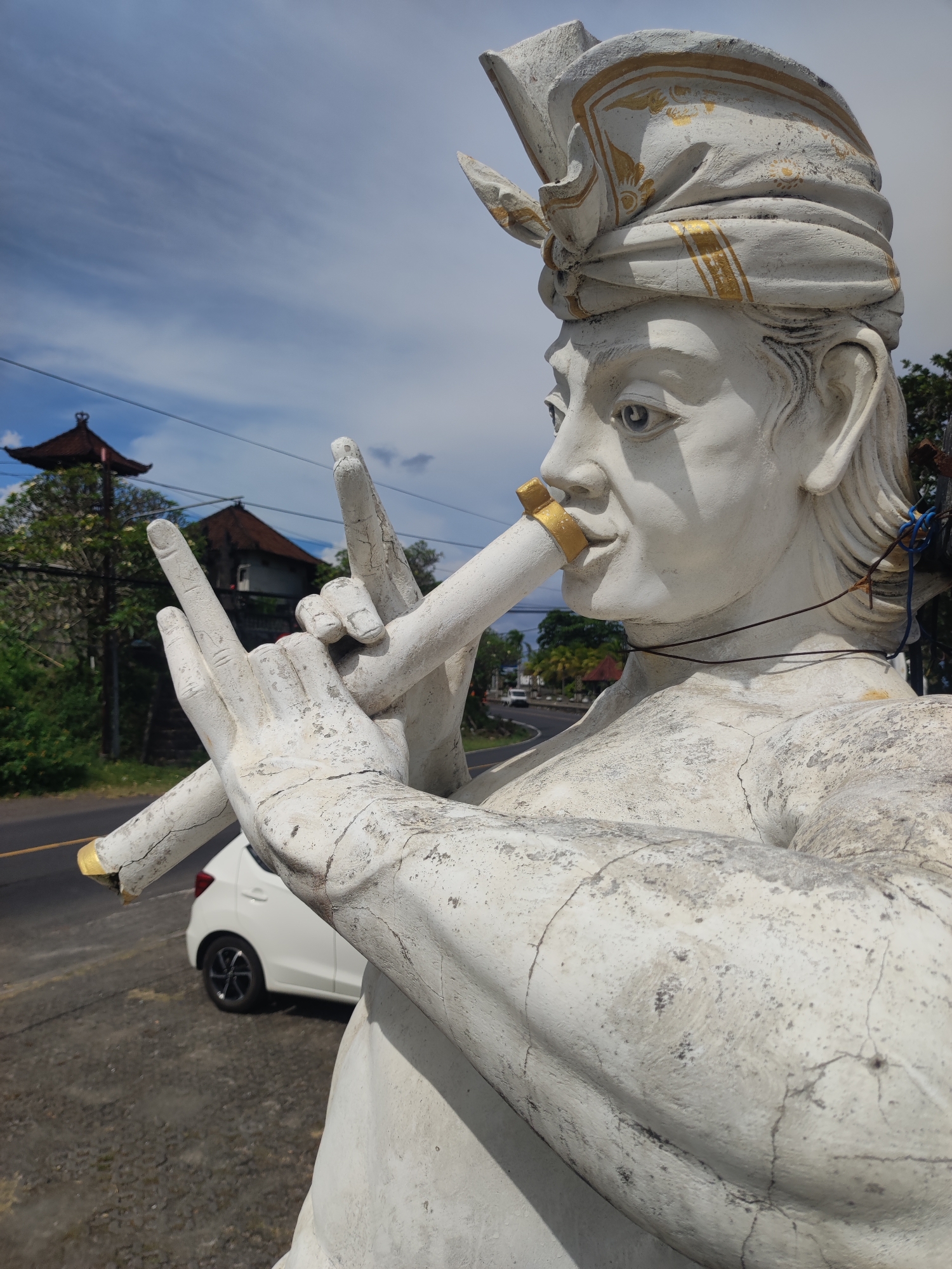 A close-up, medium shot shows a weathered, white stone statue of a male figure with a headdress, positioned roadside in Unknown, Indonesia. The figure appears to be playing a flute, with its hands positioned over a white tube with gold accents at both ends and at its mouth. Behind the statue, a white car is parked on the asphalt. A road stretches into the background with sparse traffic. The background includes buildings with red roofs and green trees, and a partly cloudy blue sky. Power lines are visible in the background. The statue shows signs of wear and tear, with cracks visible on its surface.