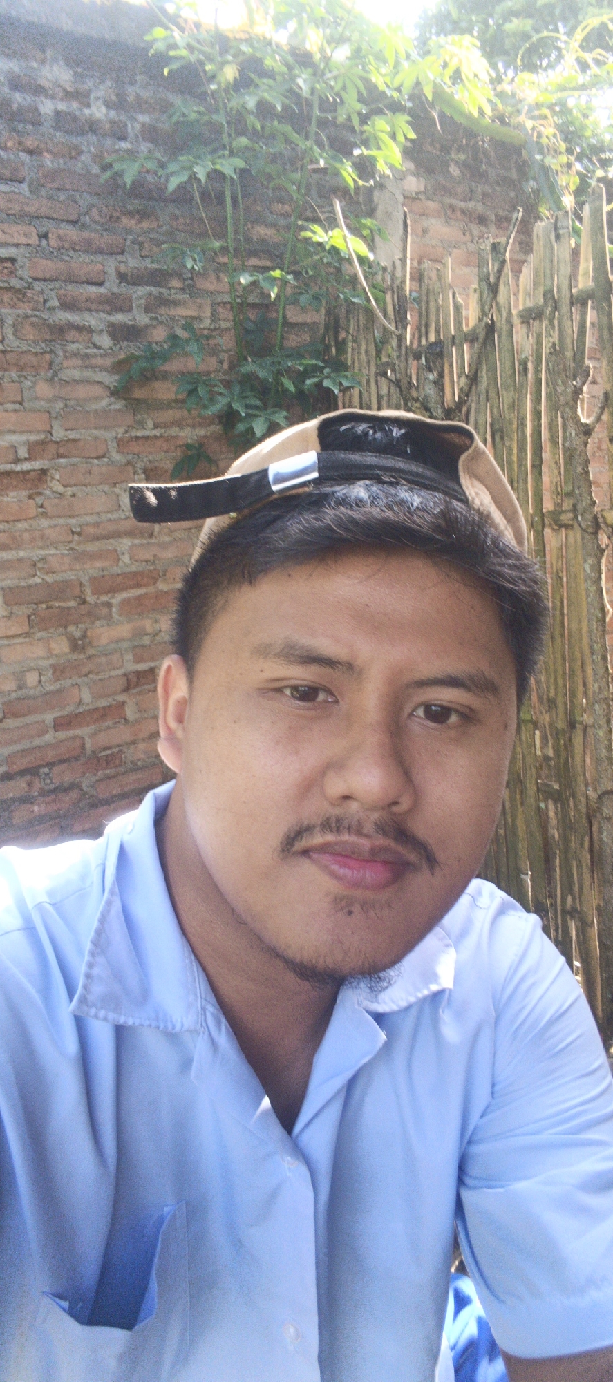 A close-up, outdoor, medium-shot photograph captures a young man in the city of Kemuning, Indonesia. He is wearing a light blue collared shirt, and a tan baseball cap is tilted backward on his head. The man has dark hair and a mustache. His expression is neutral as he looks towards the camera. The background consists of a brick wall and a wooden fence, with greenery visible above the wall. The scene is lit by natural sunlight, suggesting it is daytime.