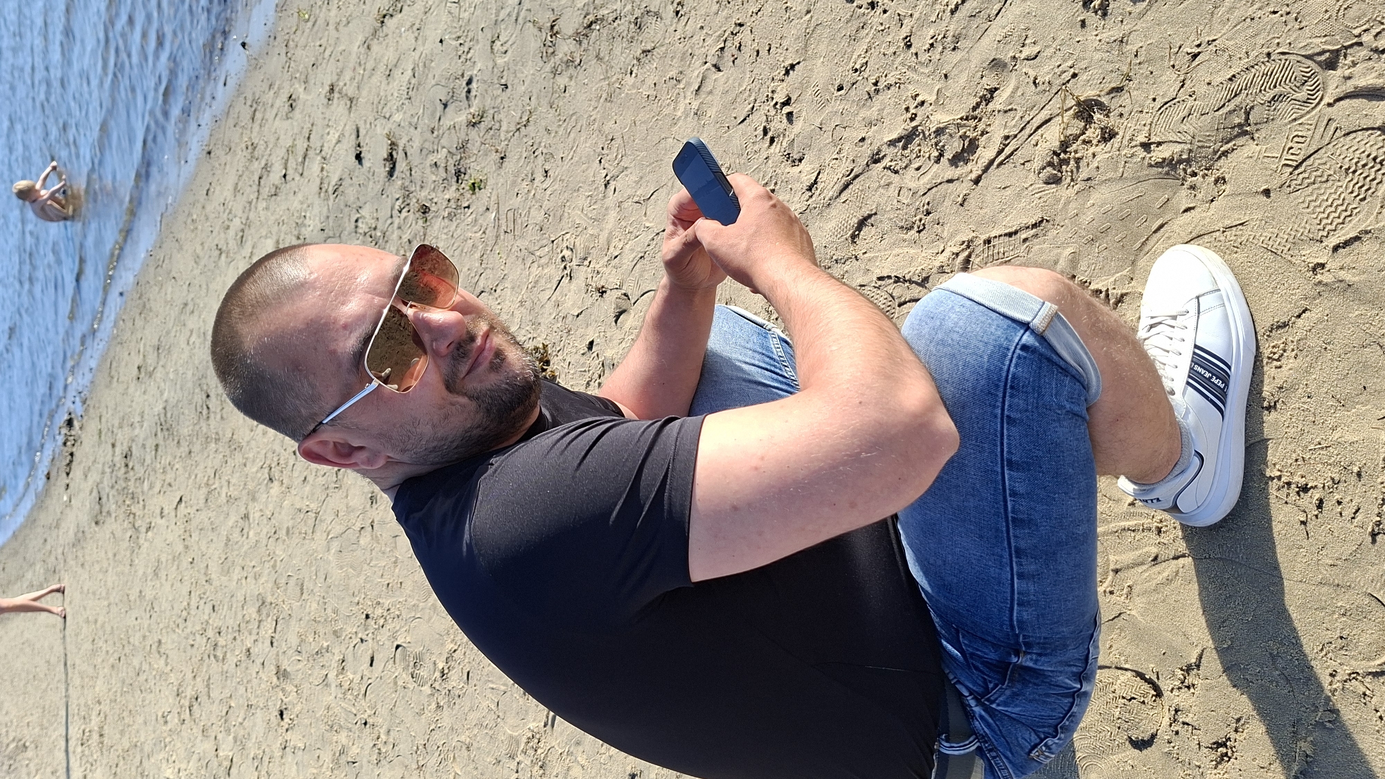 A man wearing sunglasses, a black t-shirt, and denim shorts squats on a sandy beach. He is holding a dark blue mobile phone in his right hand. White sneakers with blue stripes are on his feet. In the background, the water of a lake or sea is visible, with a child playing in the shallow water. In the far background, a person's legs and feet are visible on the sand. The lighting suggests it is daytime, and the scene appears to be a public recreational area. This location is identified as a beach in Tallinn, Estonia.